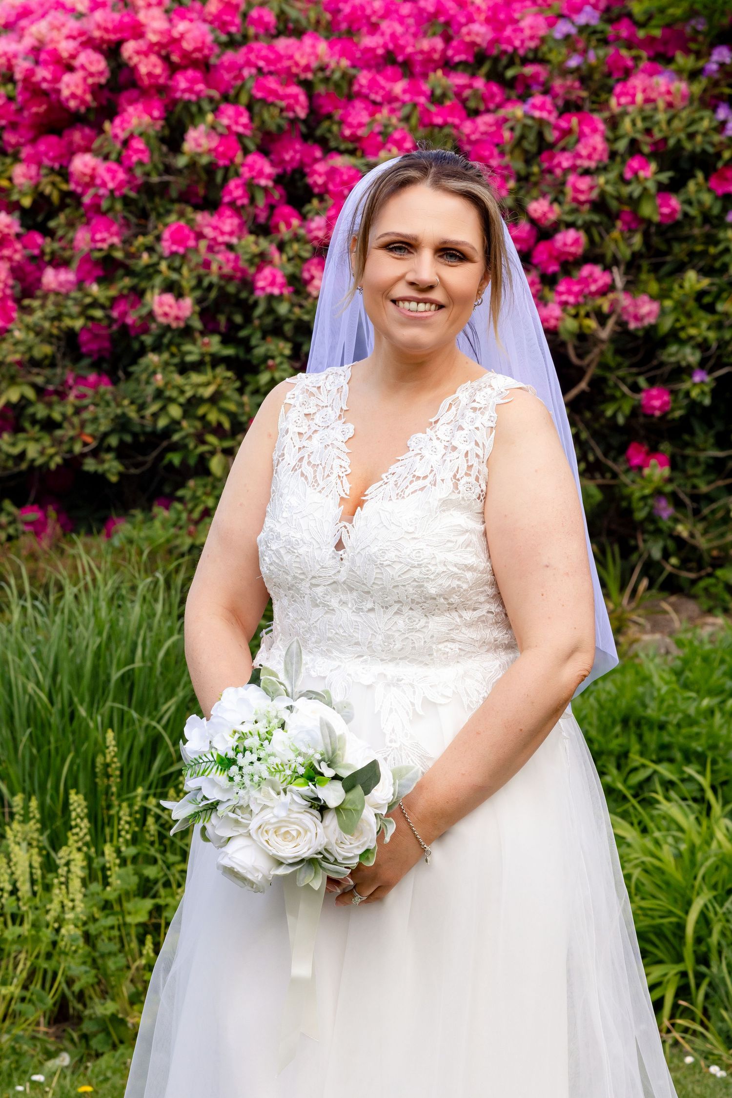 Smiling bride in white lace gown and veil holds white bouquet before vibrant pink flowering shrubs.