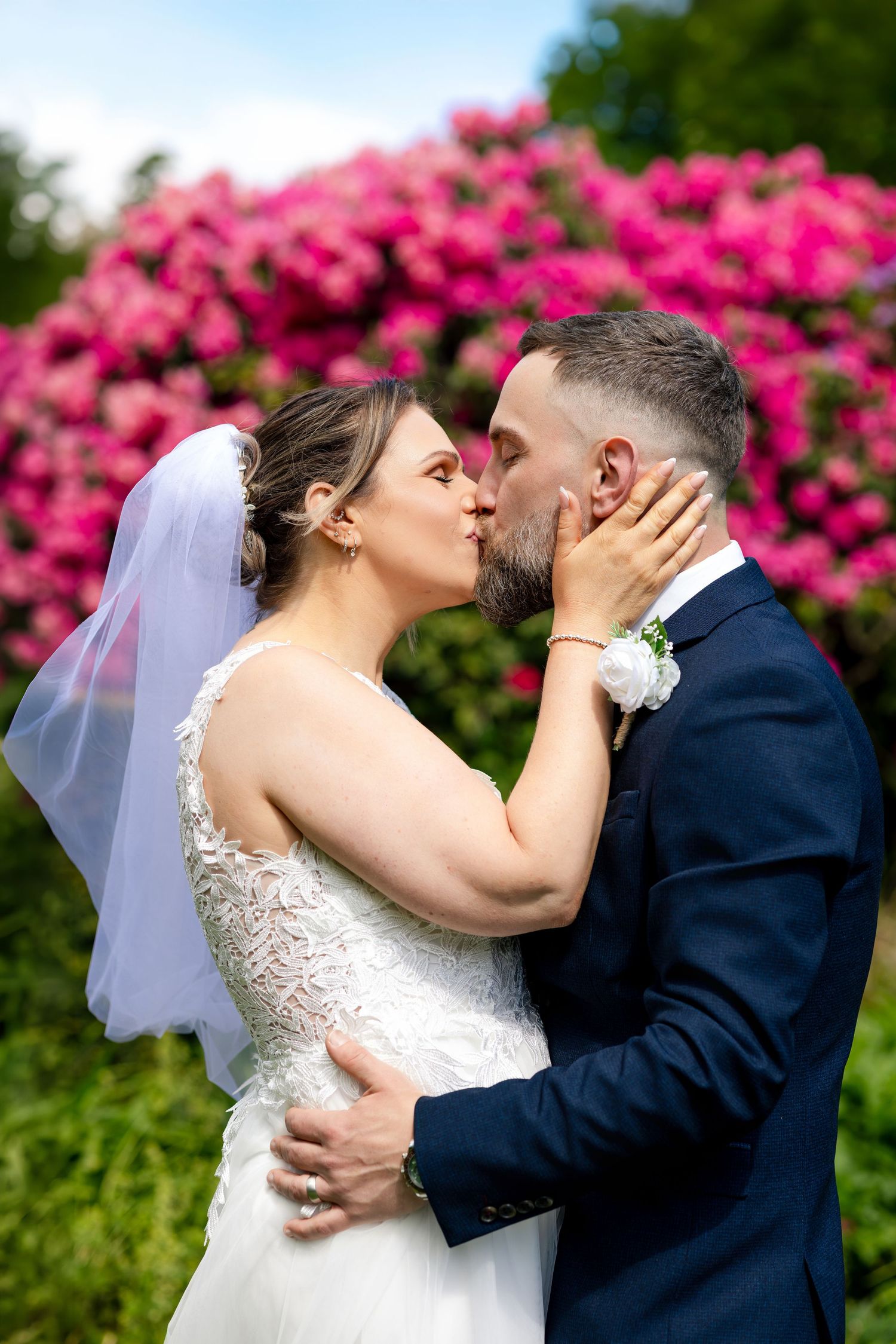 Bride and groom sharing a romantic kiss in front of vibrant pink flowering bushes on their wedding day.