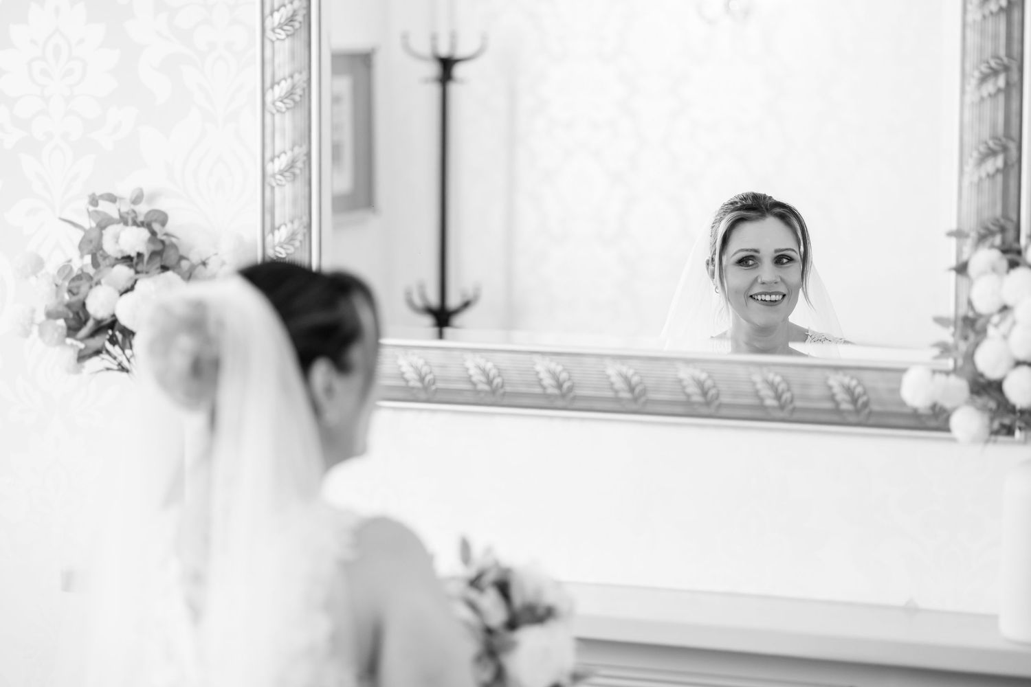 Bride in wedding dress and veil smiles at her reflection in an ornate mirror, captured in black and white.