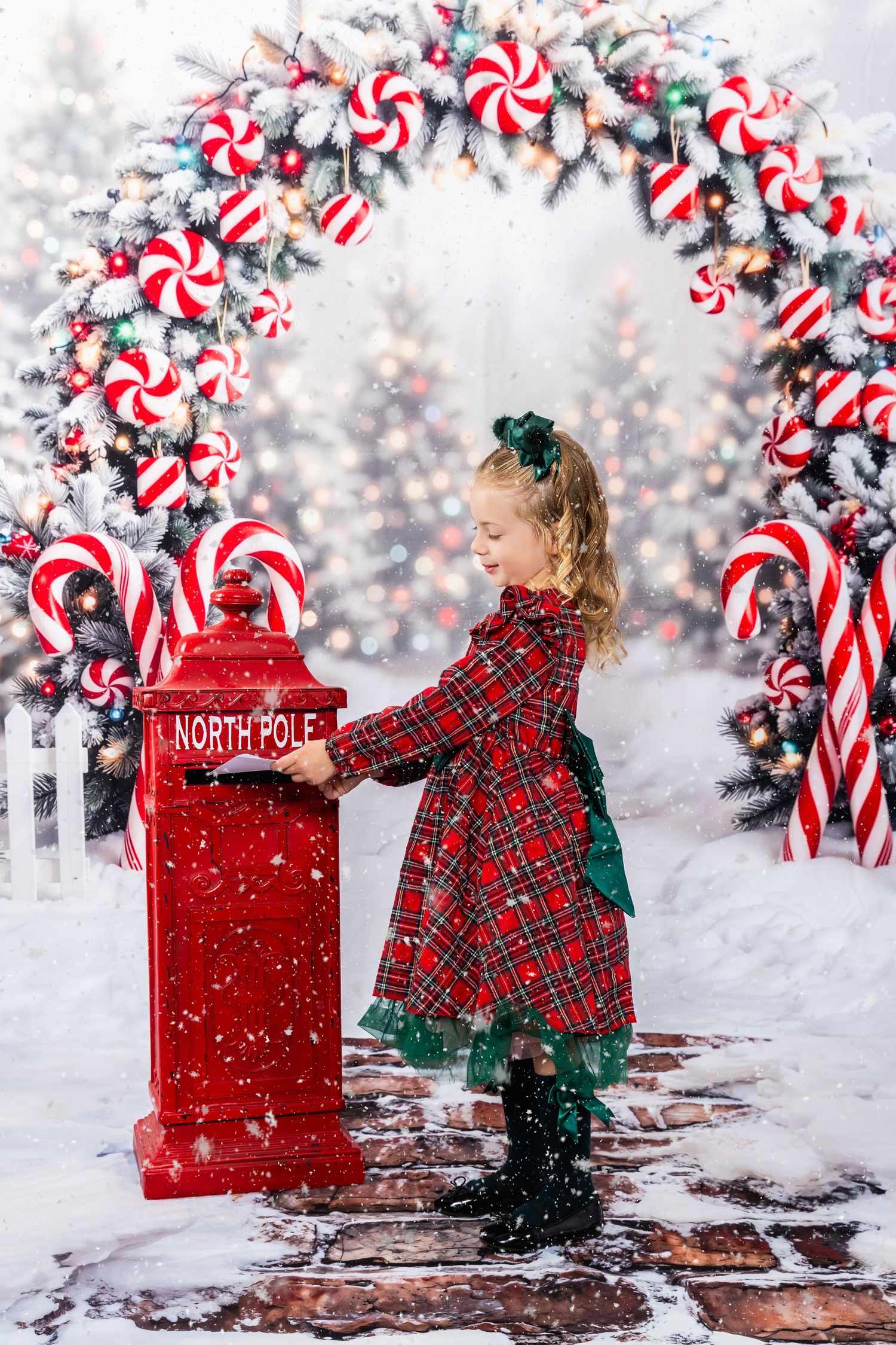 Girl in plaid dress mailing letter to Santa at red North Pole mailbox with candy cane arch backdrop.