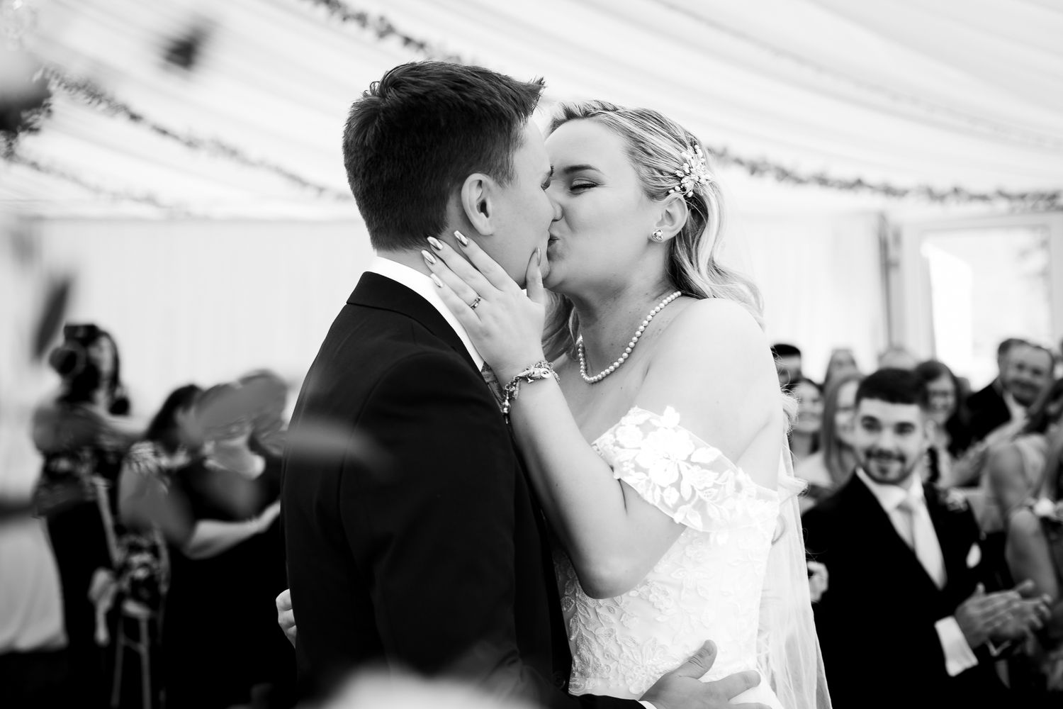 Bride and groom share their first kiss surrounded by celebrating guests in a black and white reception photo.