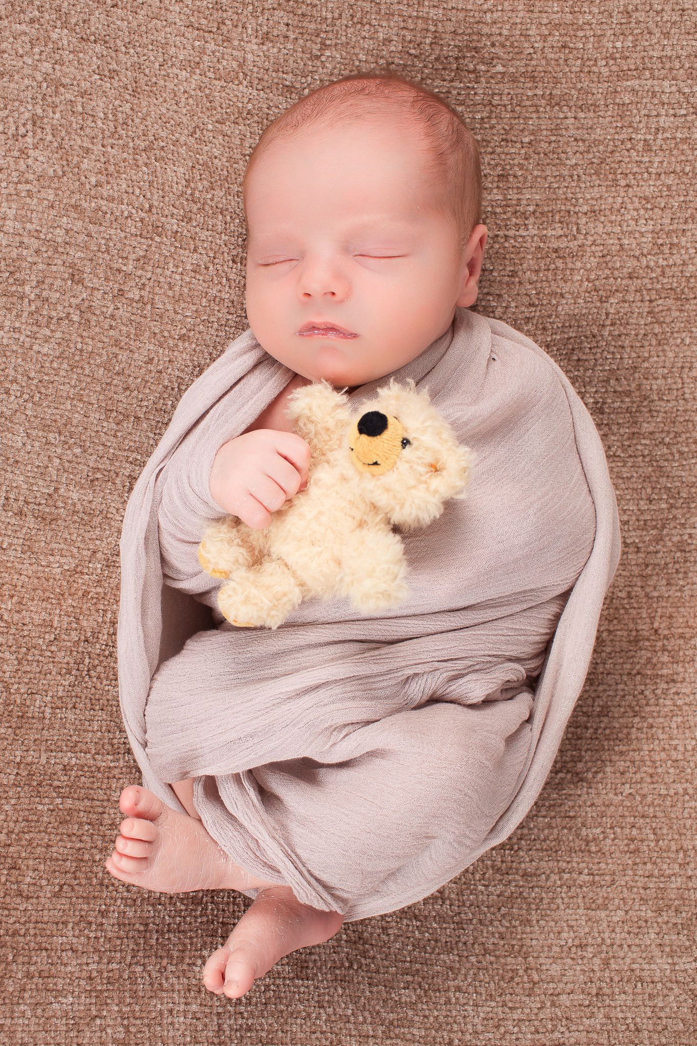 Newborn baby sleeping peacefully in a gray swaddle while holding a small teddy bear.