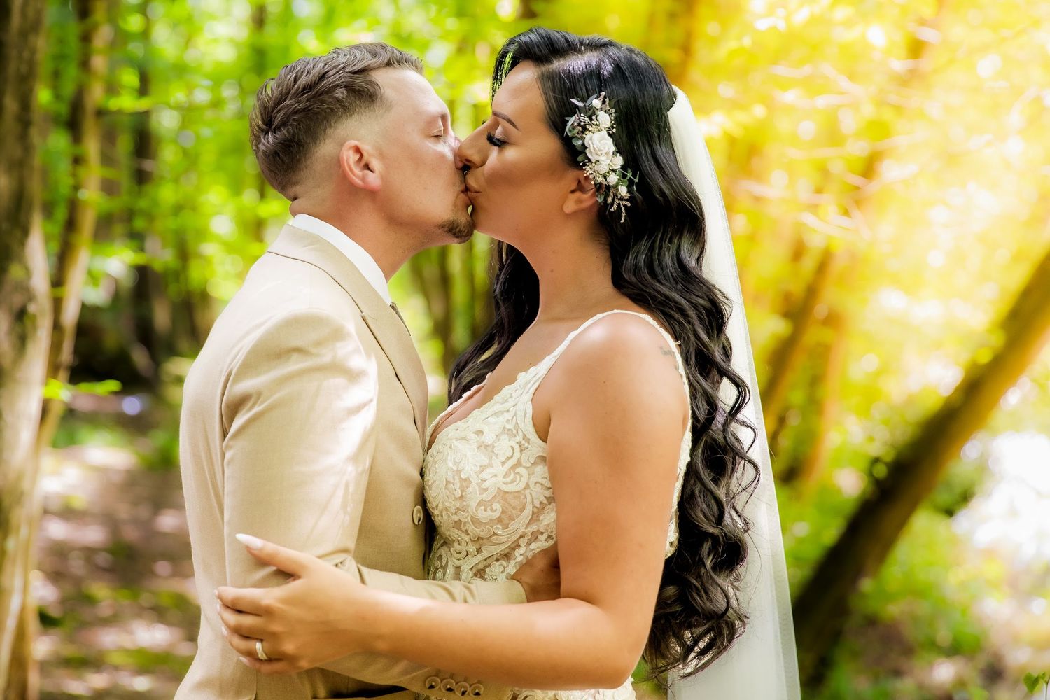 Bride and groom kissing in a sunlit forest, bride in a lace gown with flowers in her hair.