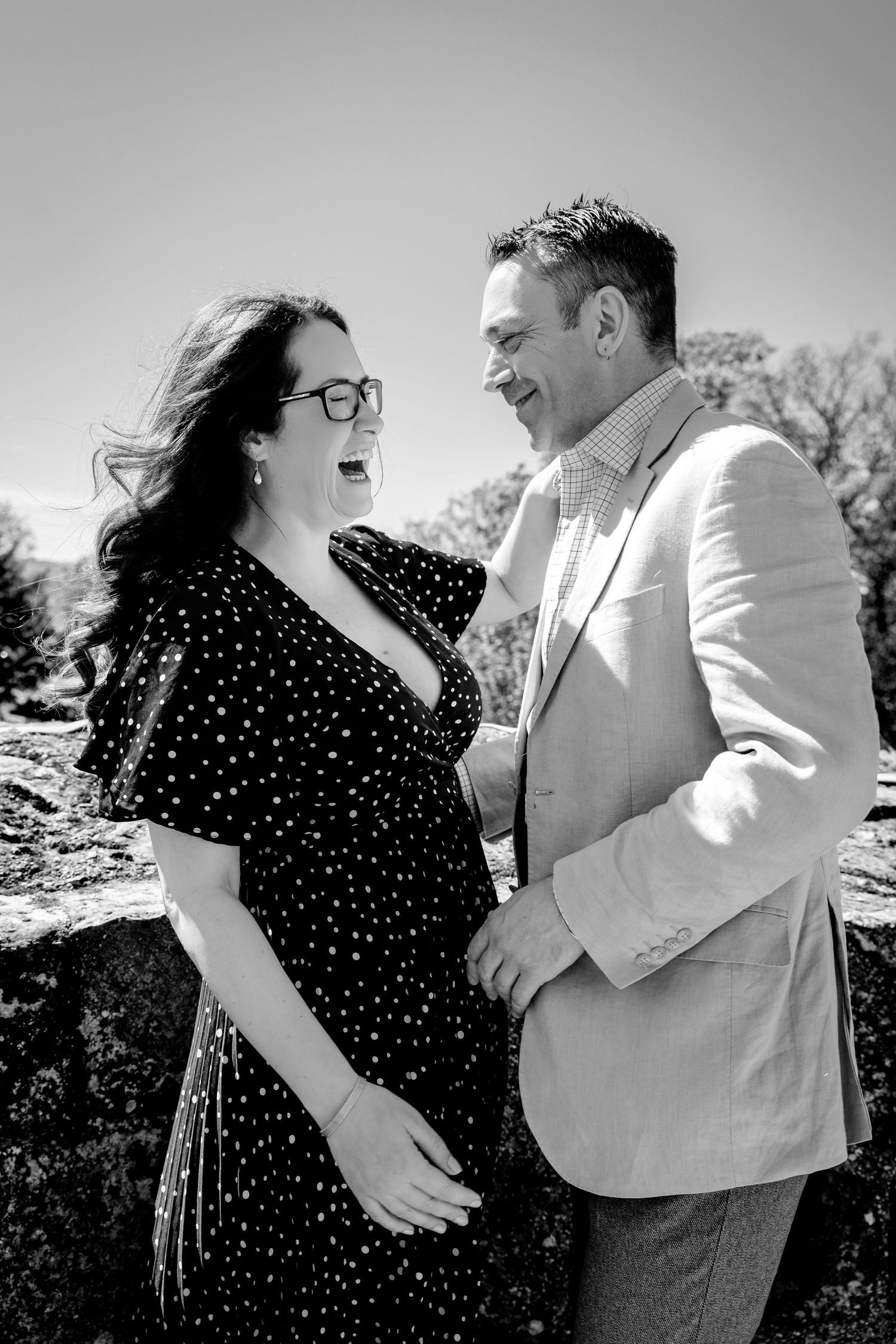 Black and white photo of a laughing couple outdoors, woman in polka dot dress and glasses, man in light blazer.