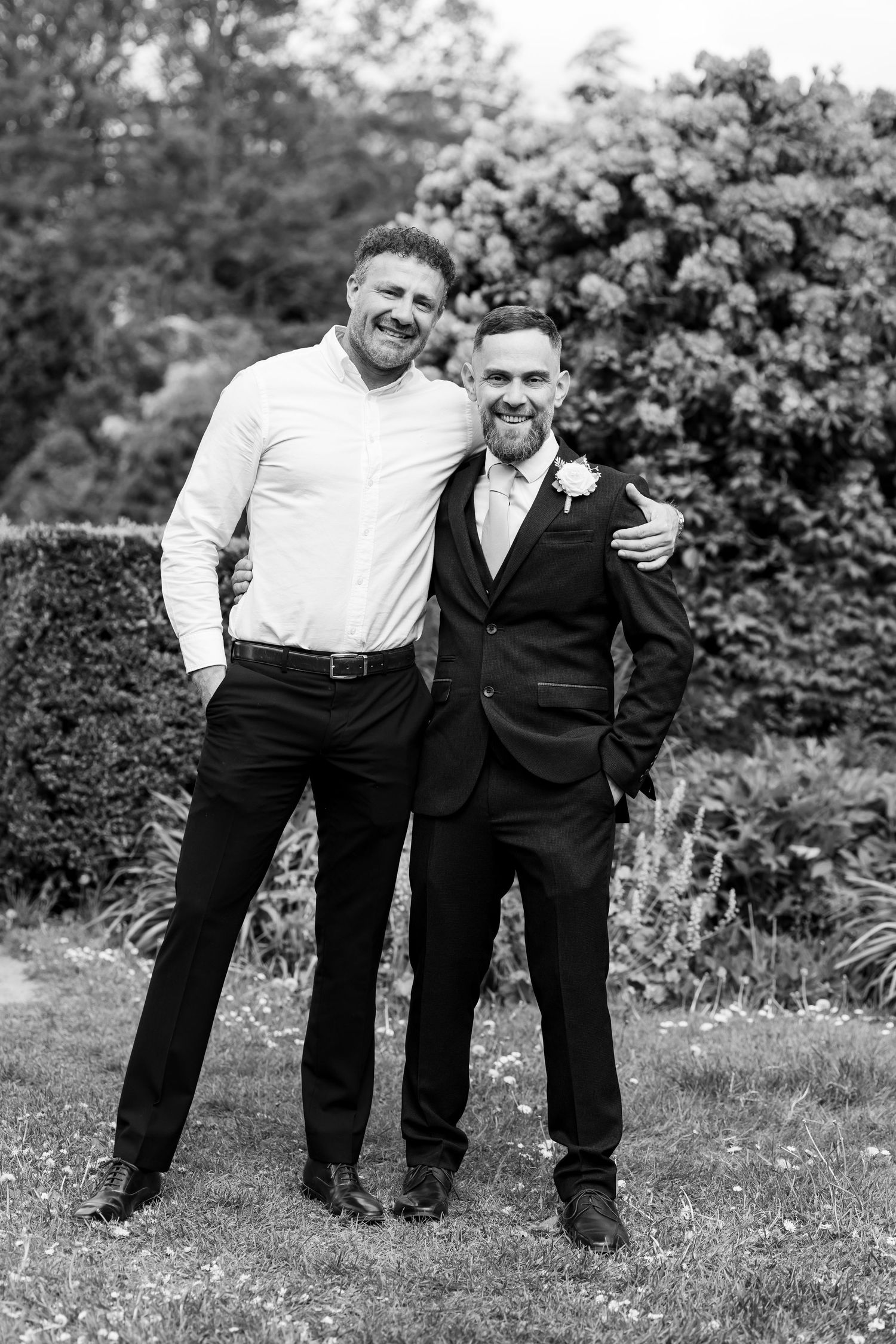 Two men in formal attire smiling together outdoors at a wedding, one wearing a suit with boutonniere.