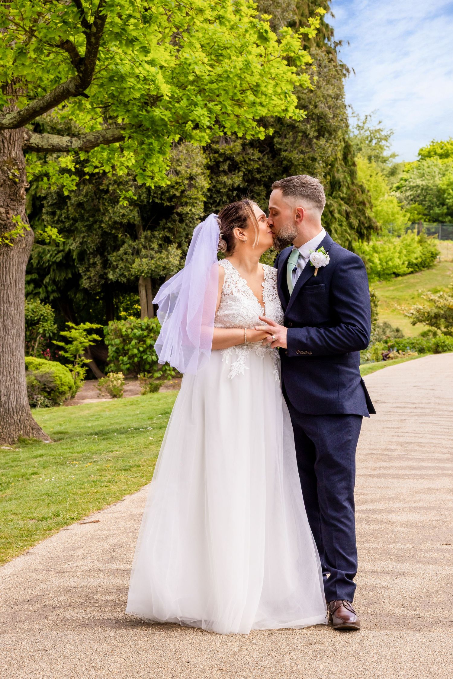 Bride and groom sharing a kiss on a garden path, bride in white lace gown with veil, groom in navy suit.