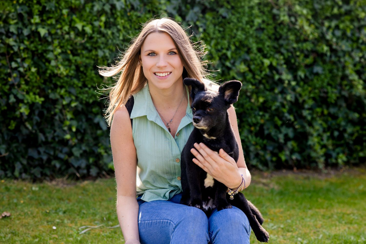 A smiling blonde woman crouches on grass holding a small black dog in front of green hedges.
