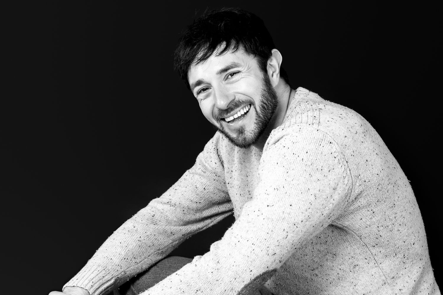 A smiling bearded man in a speckled sweater poses in a casual black and white studio portrait.
