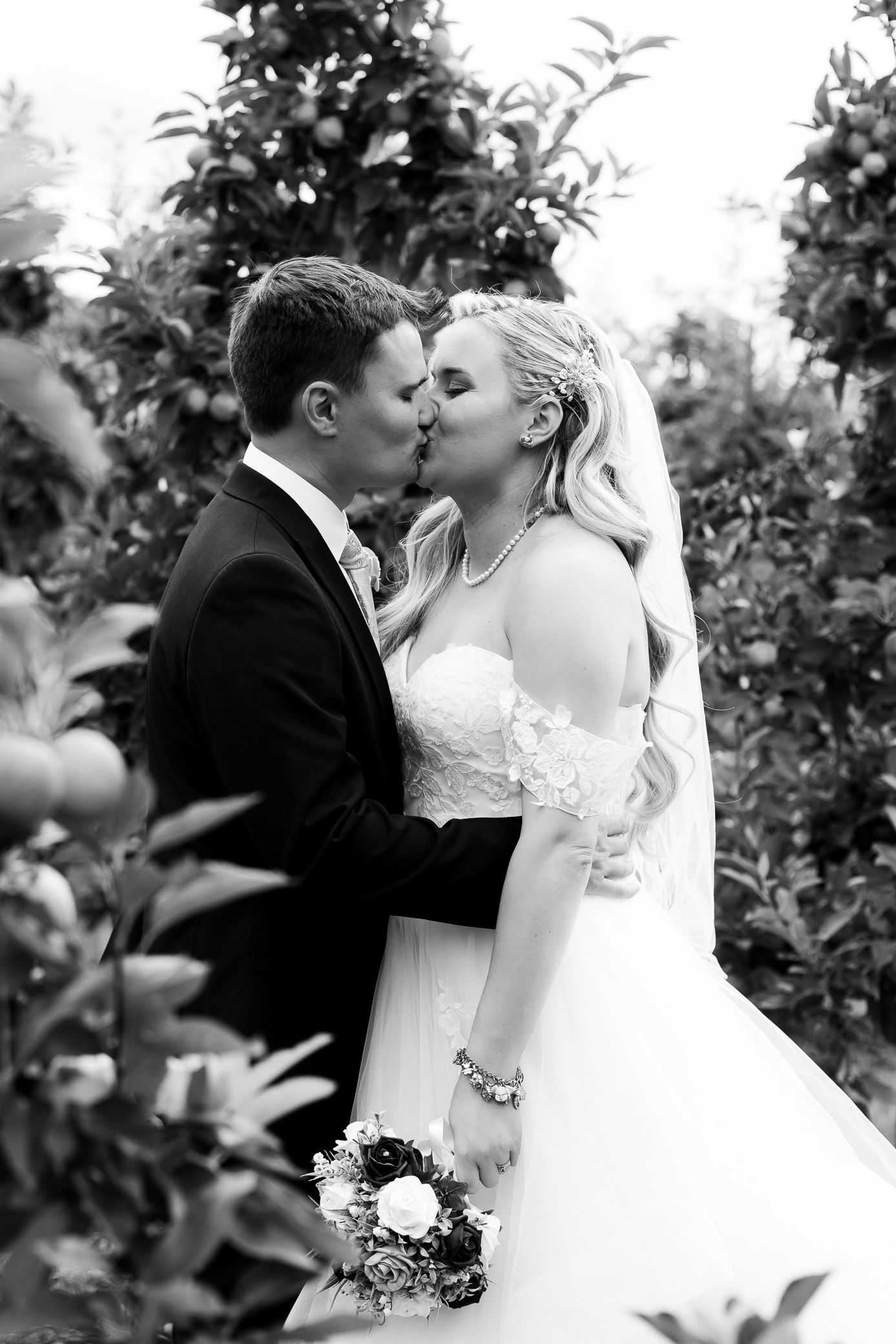 Bride and groom share a romantic kiss surrounded by lush greenery in a black and white portrait.