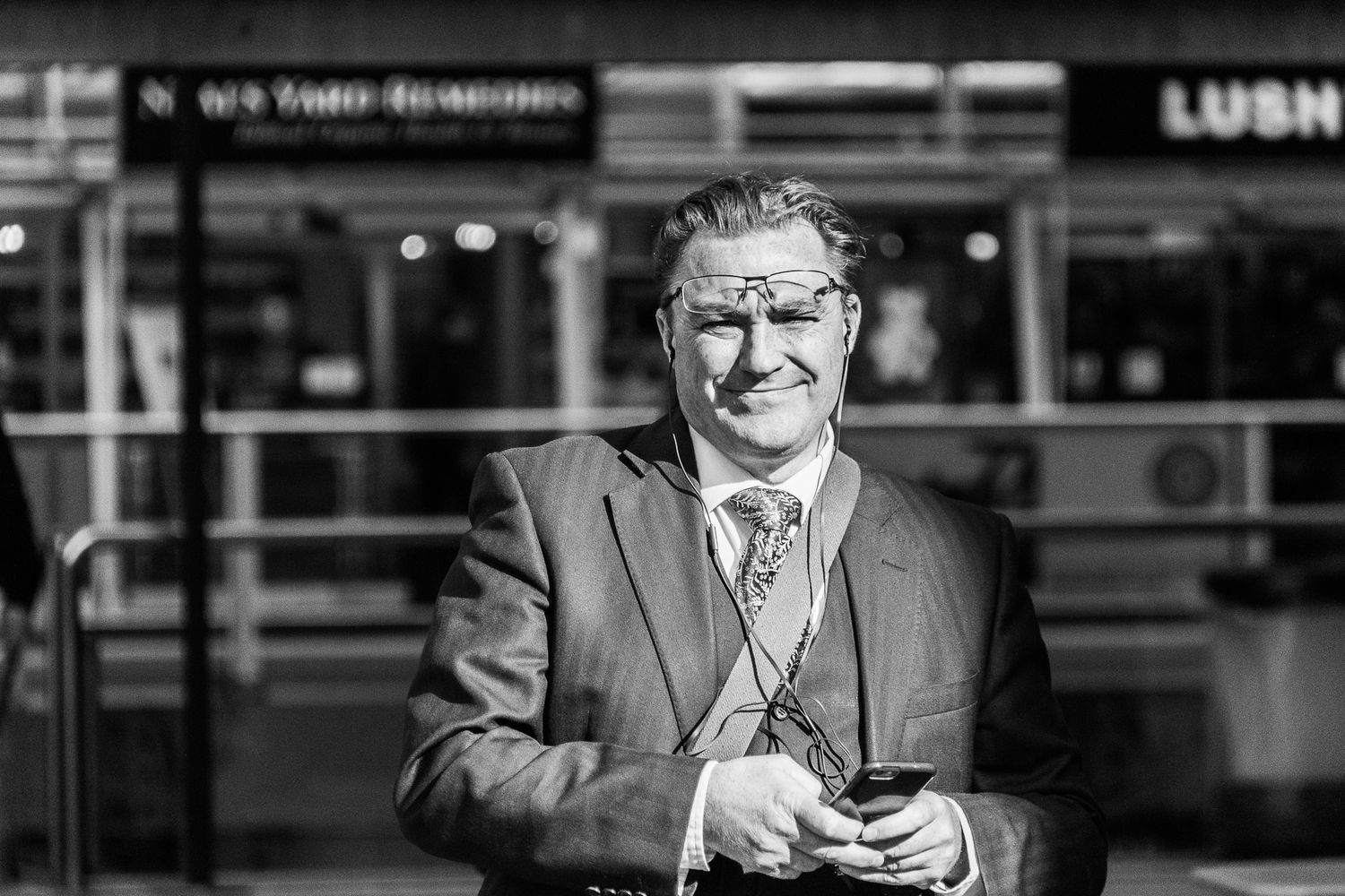 Smiling middle-aged businessman in suit holding phone outside a store in black and white photo.