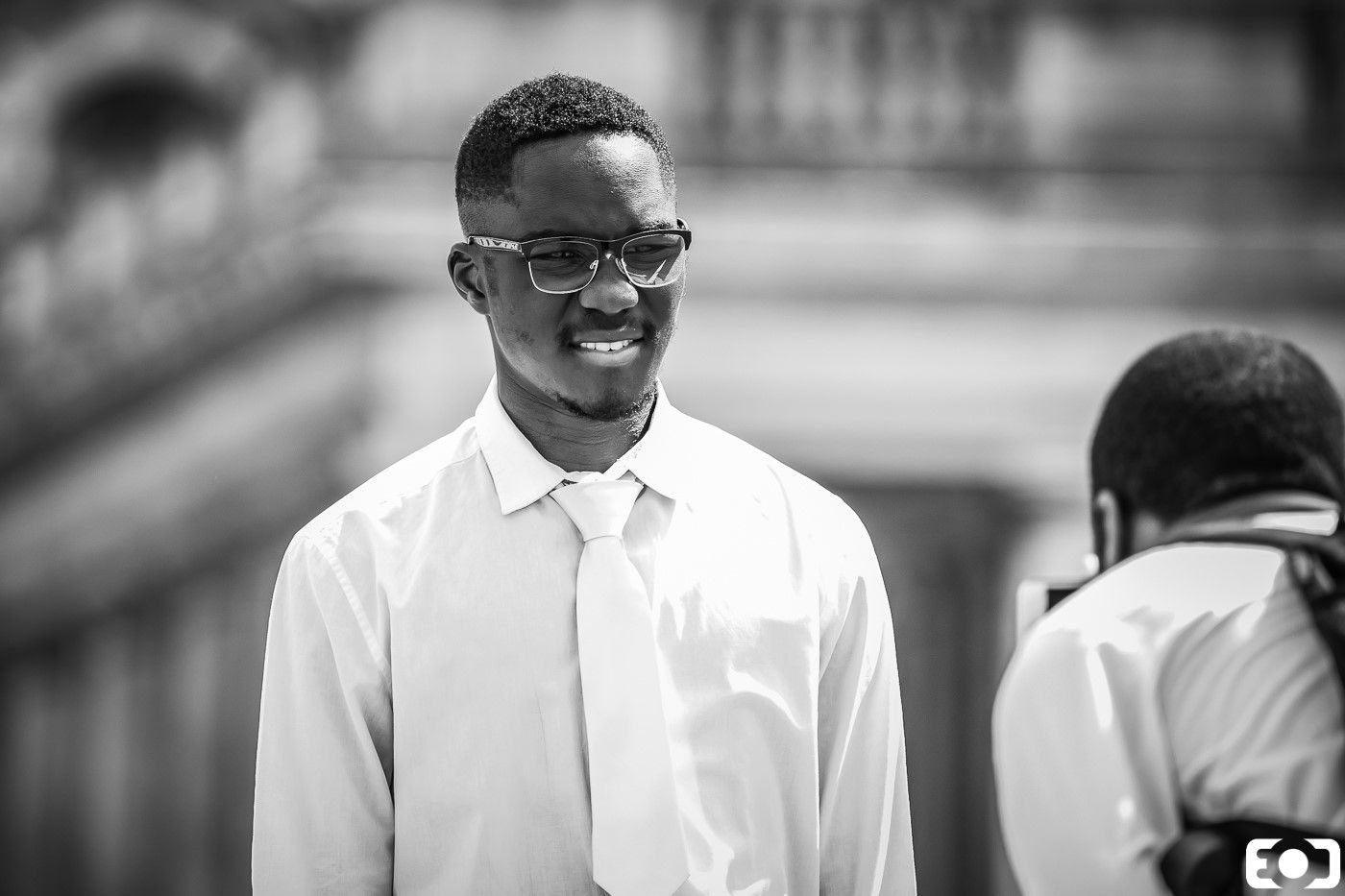 Smiling young man in white shirt and tie, candid black and white portrait with architectural background.