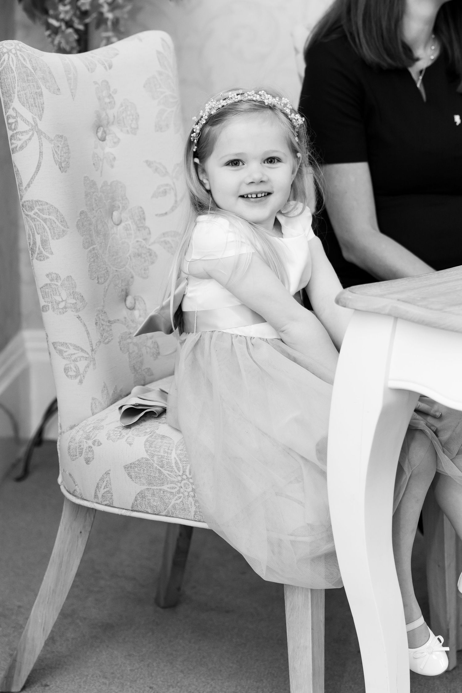 A smiling young girl in a white dress and floral crown sits on a floral chair at a white table in black and white photo.