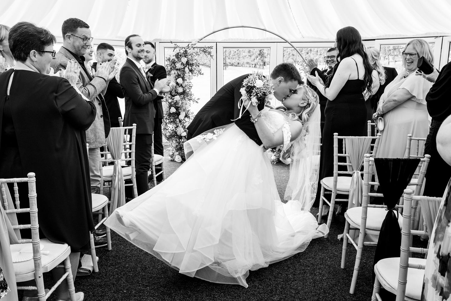 Black and white photo of bride and groom sharing a romantic dip kiss at their wedding ceremony aisle.
