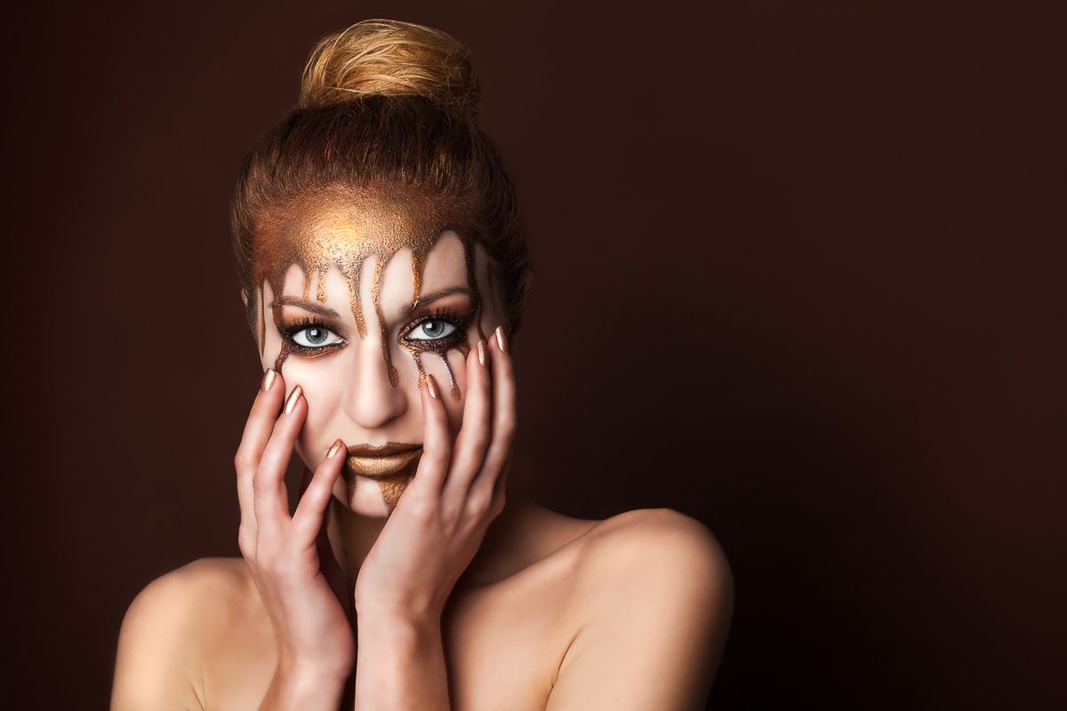 A woman with dramatic golden face paint holds her face with both hands against a dark background.