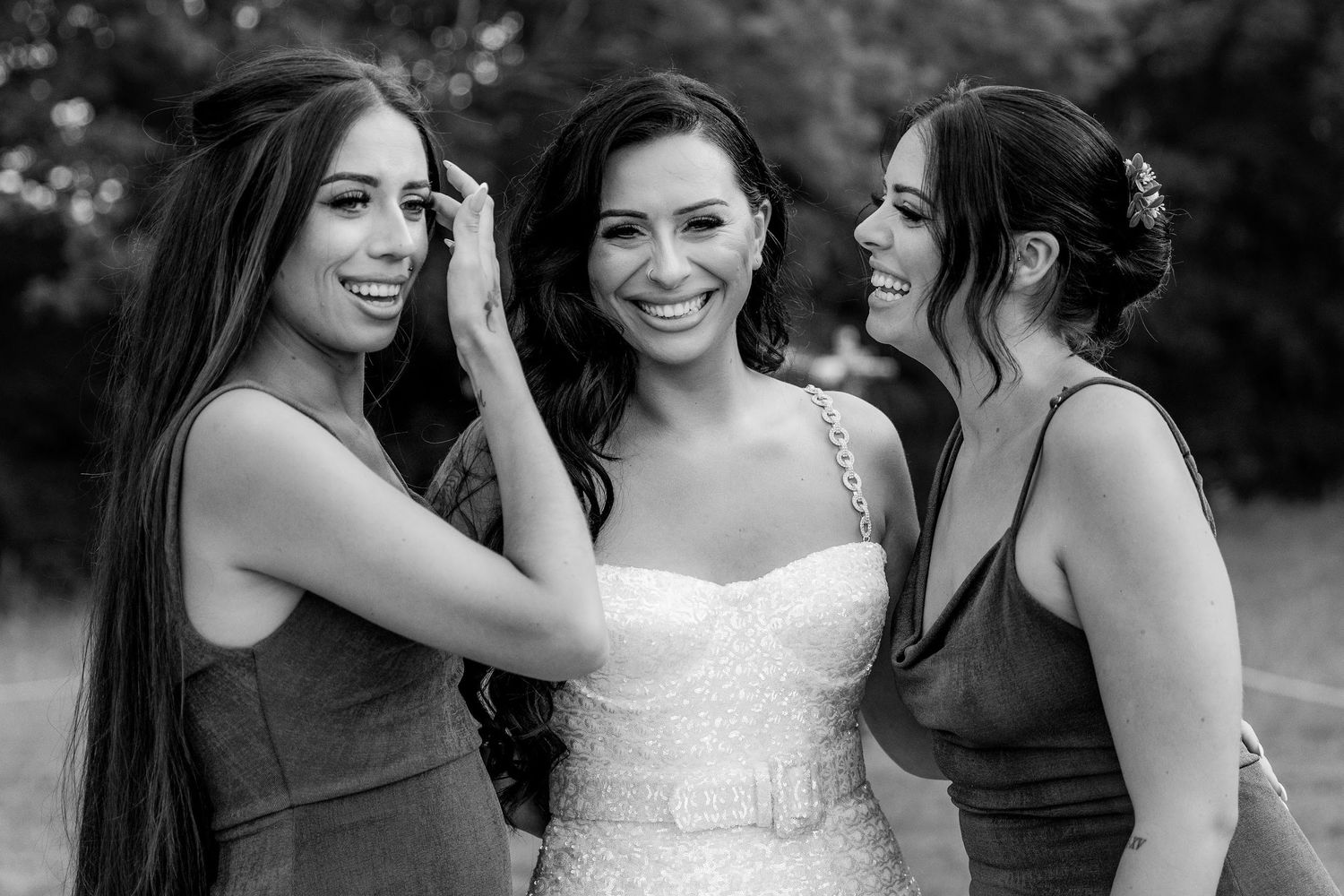 Black and white photo of a bride with two bridesmaids laughing and fixing her hair outdoors.