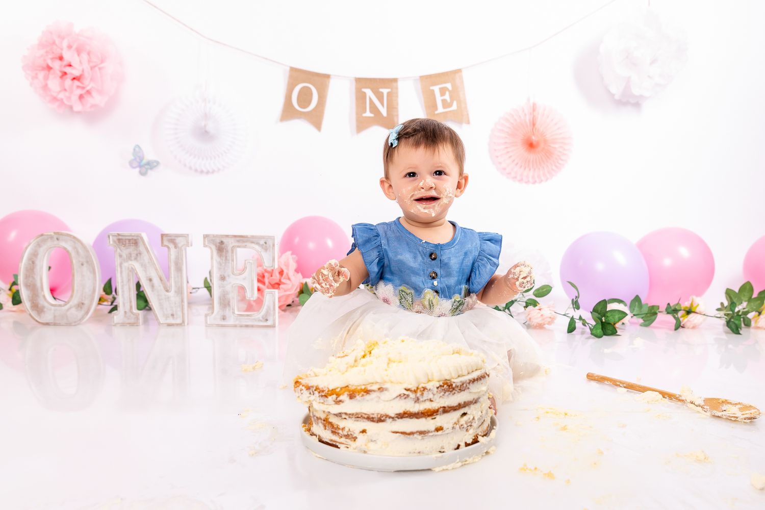 Smiling baby girl celebrating first birthday smash cake session with pink balloons and ONE banner.