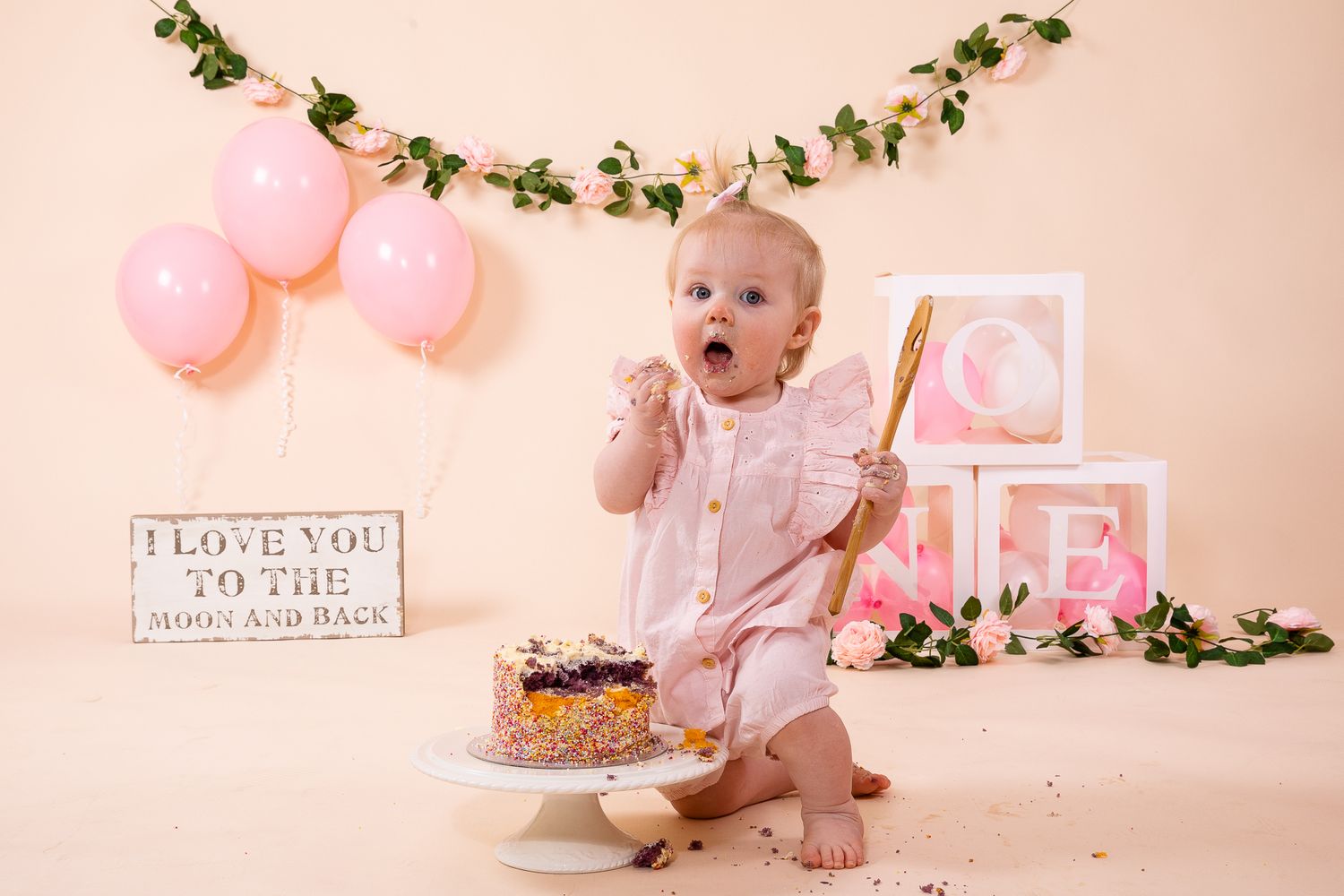 Baby girl in pink dress celebrates first birthday with cake smash, surrounded by balloons and ONE blocks.