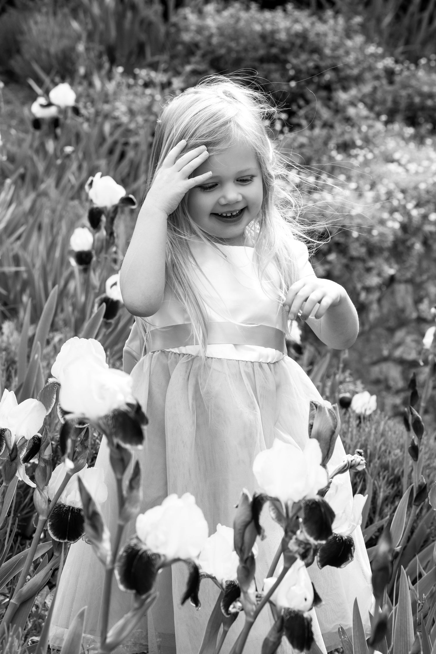 Black and white photo of a laughing blonde girl in a dress standing among blooming iris flowers in a garden.