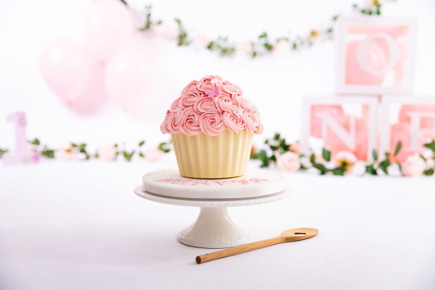 A large pink rosette frosted cupcake on a white cake stand with a wooden spoon, pink balloons and letter blocks in background.