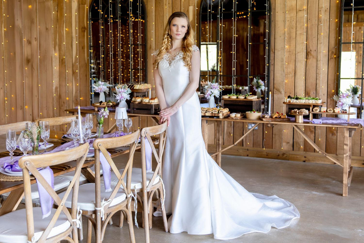 Elegant bride in satin wedding gown standing in a rustic barn venue with fairy light decor.