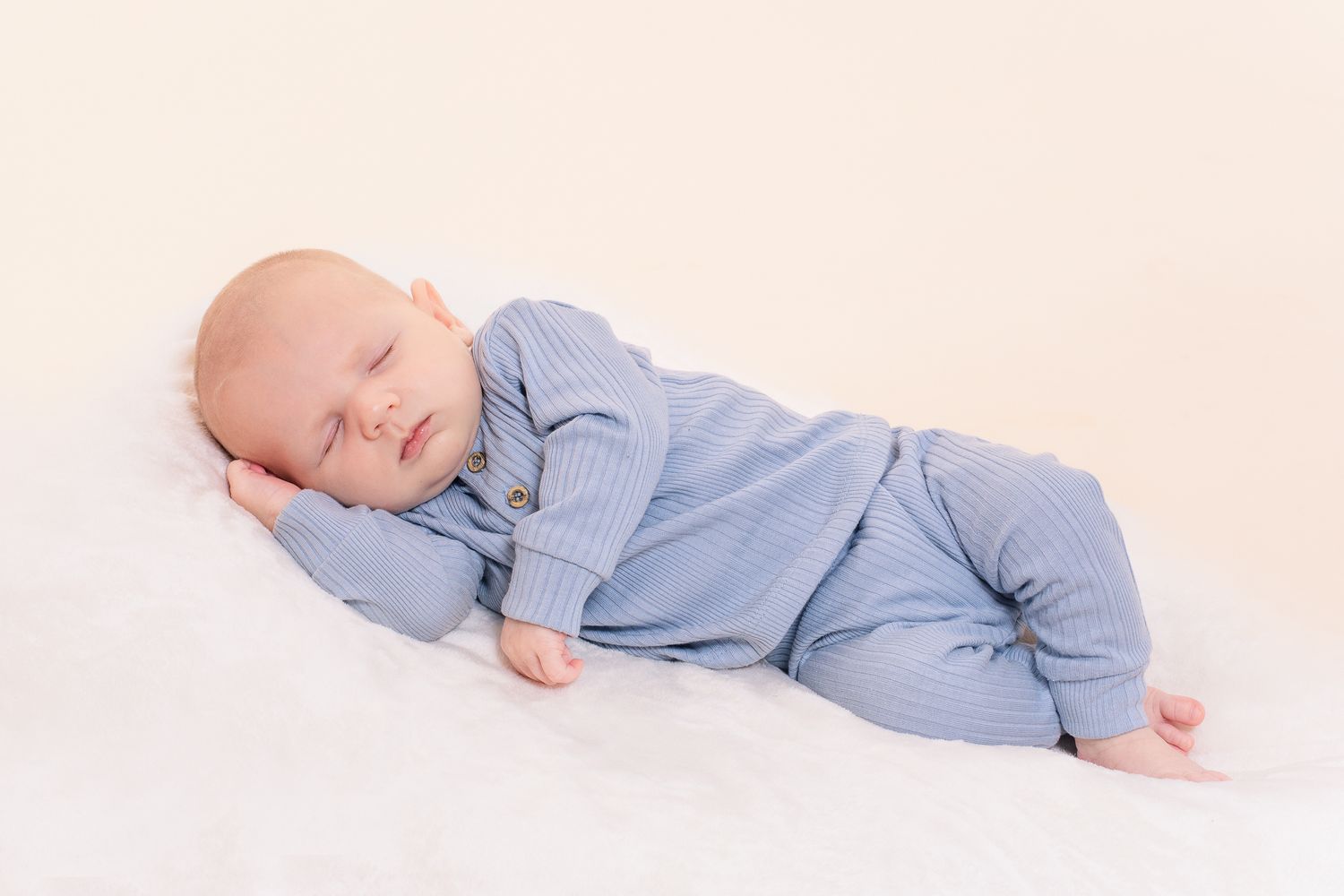 Newborn baby sleeping soundly in a blue ribbed outfit lying on a soft white blanket in studio lighting.
