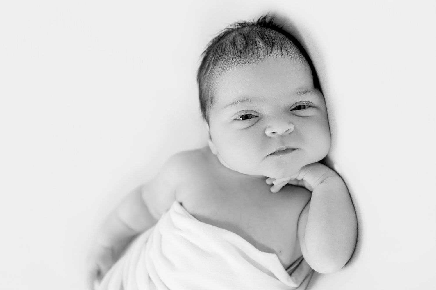 Awake newborn baby wrapped in white, resting chin on hand, black and white portrait photo.