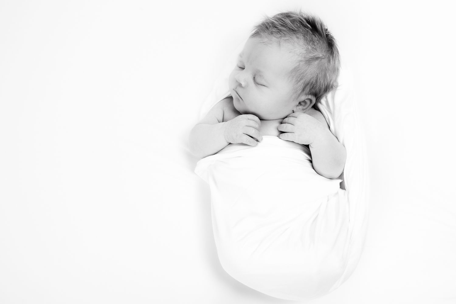 Black and white newborn portrait of a sleeping baby swaddled in white fabric with hands near face.