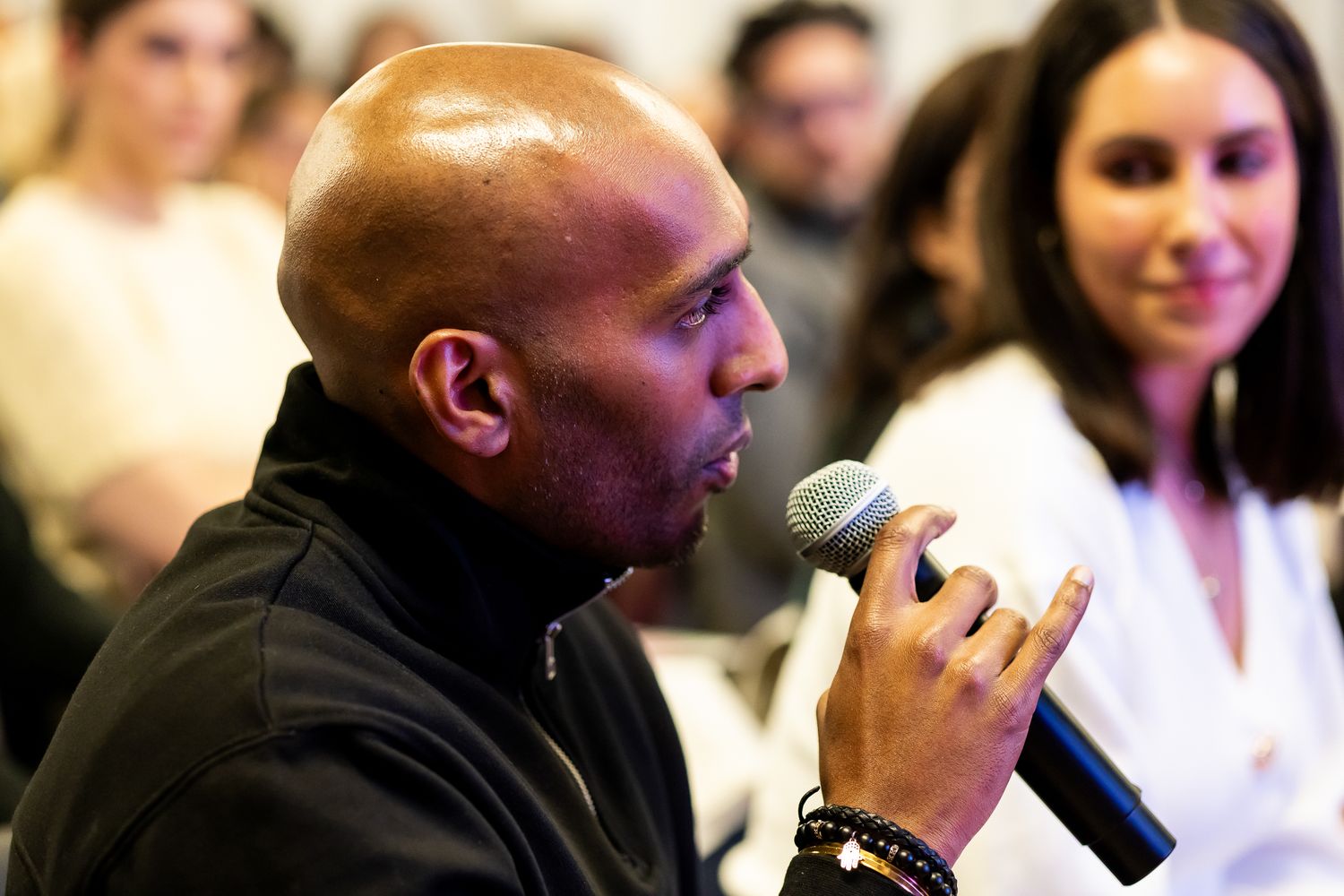 Bald man speaking into a microphone at an indoor event with audience in background.
