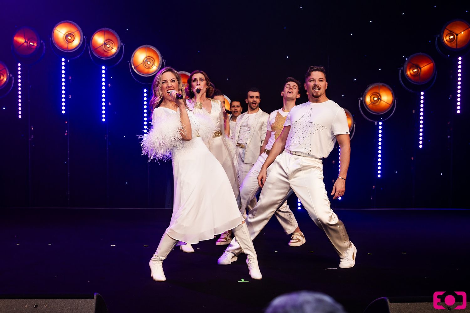 Four performers in white outfits dance on a lit stage with orange spotlights during a live show.
