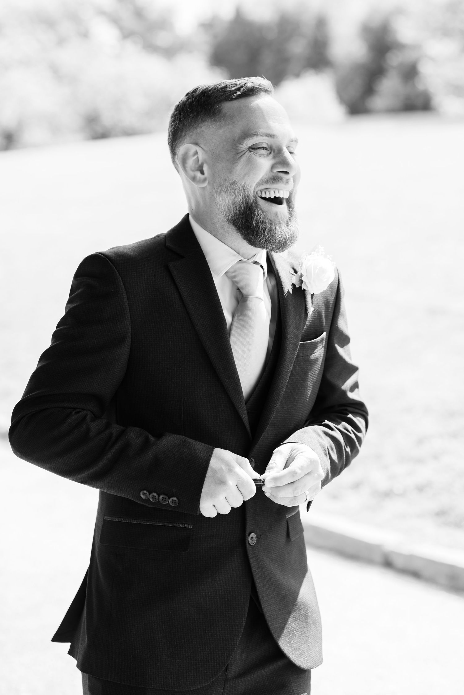 Smiling groom in black suit adjusting his jacket button outdoors on his wedding day.