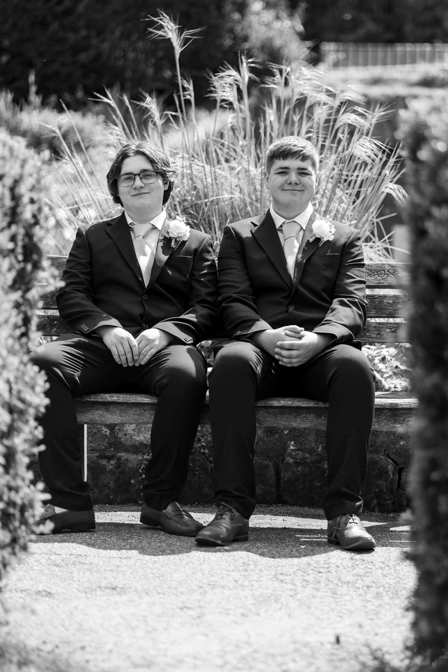 Two young men in suits with boutonnieres sit on a stone bench in a garden, black and white photo.
