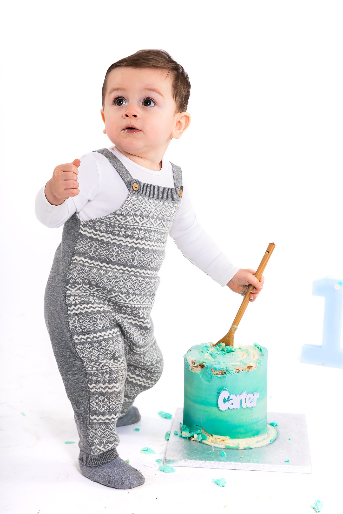 Baby boy in gray overalls holding a gold spoon next to a teal first birthday smash cake named Carter.