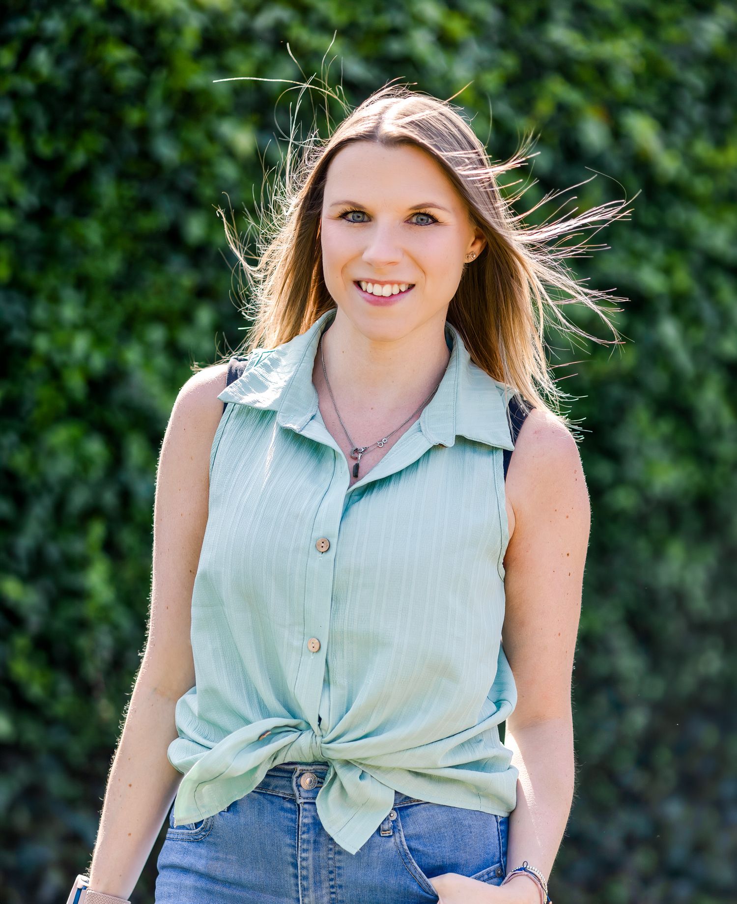 A cheerful blonde woman in a mint sleeveless top and jeans walks outdoors near a green hedge wall.