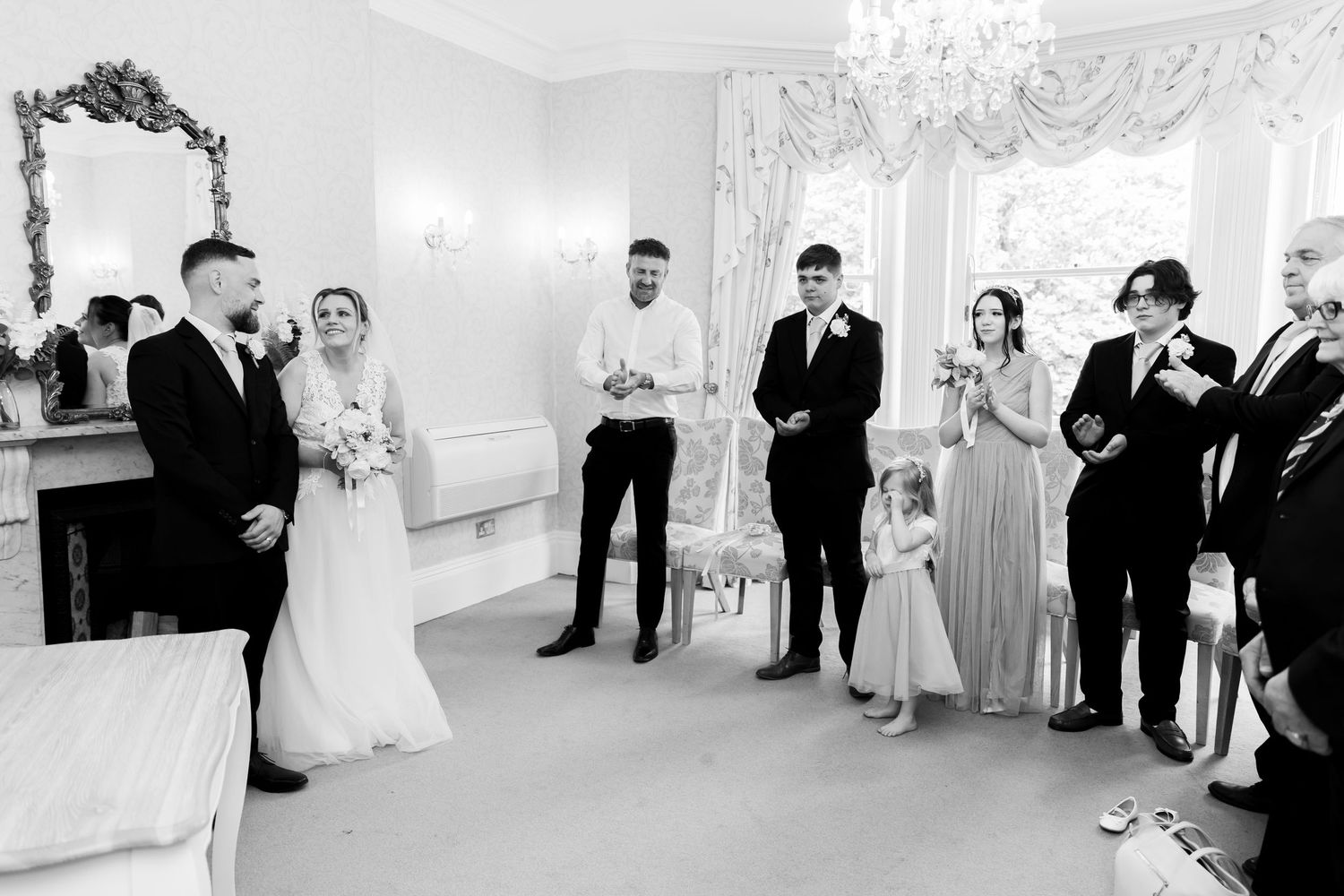 Black and white wedding ceremony photo with bride, groom, and guests gathered in an elegant room.