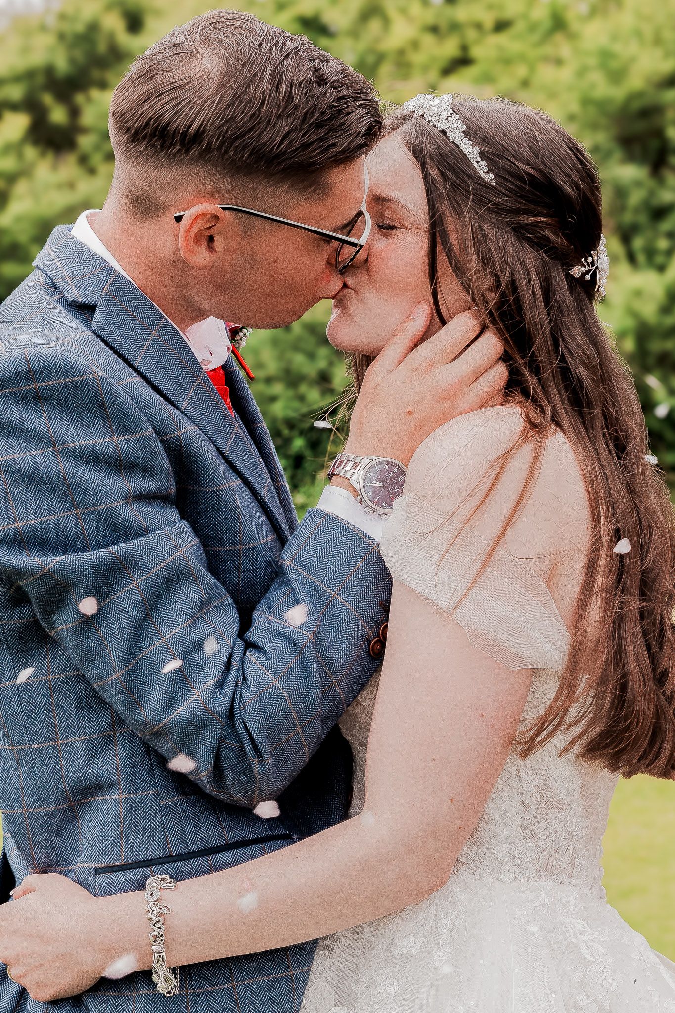 Bride and groom sharing a tender kiss outdoors, confetti falling, groom in blue tweed suit.