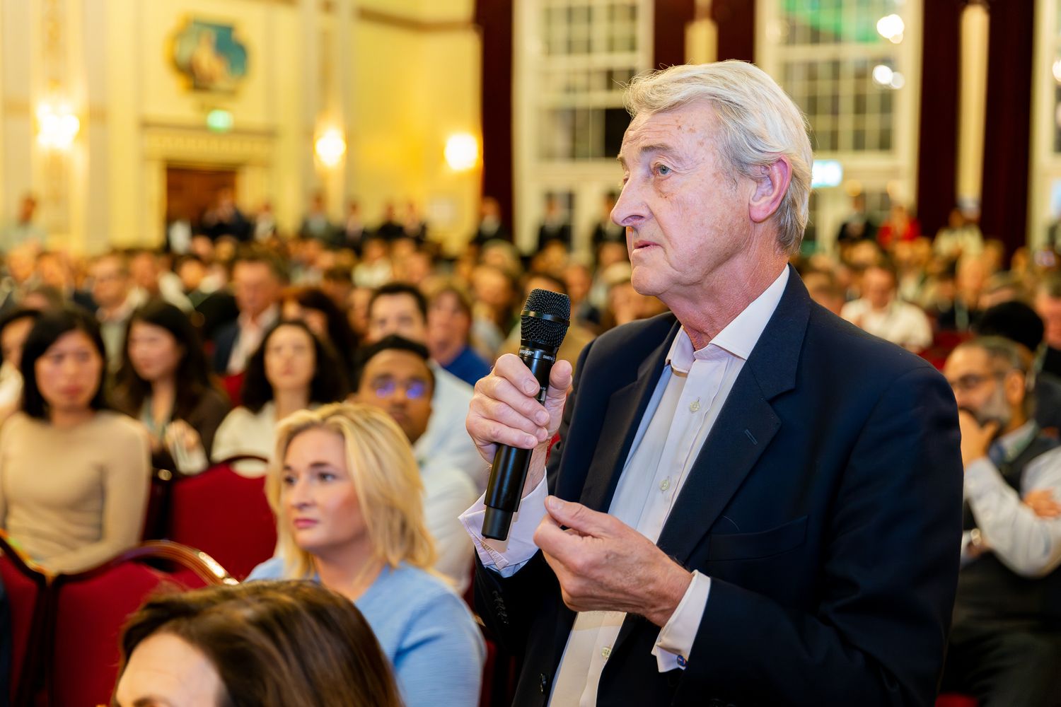 Elderly man speaking into microphone at a crowded conference hall event.