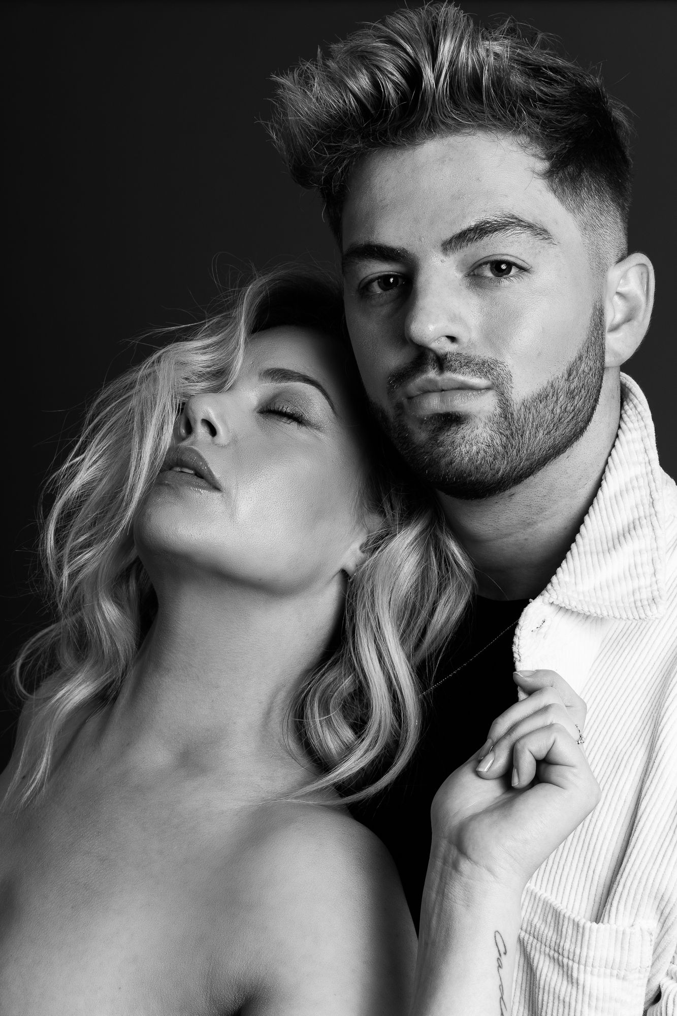 Black and white portrait of a couple, man with beard looking at camera, woman with blonde hair leaning on his shoulder.