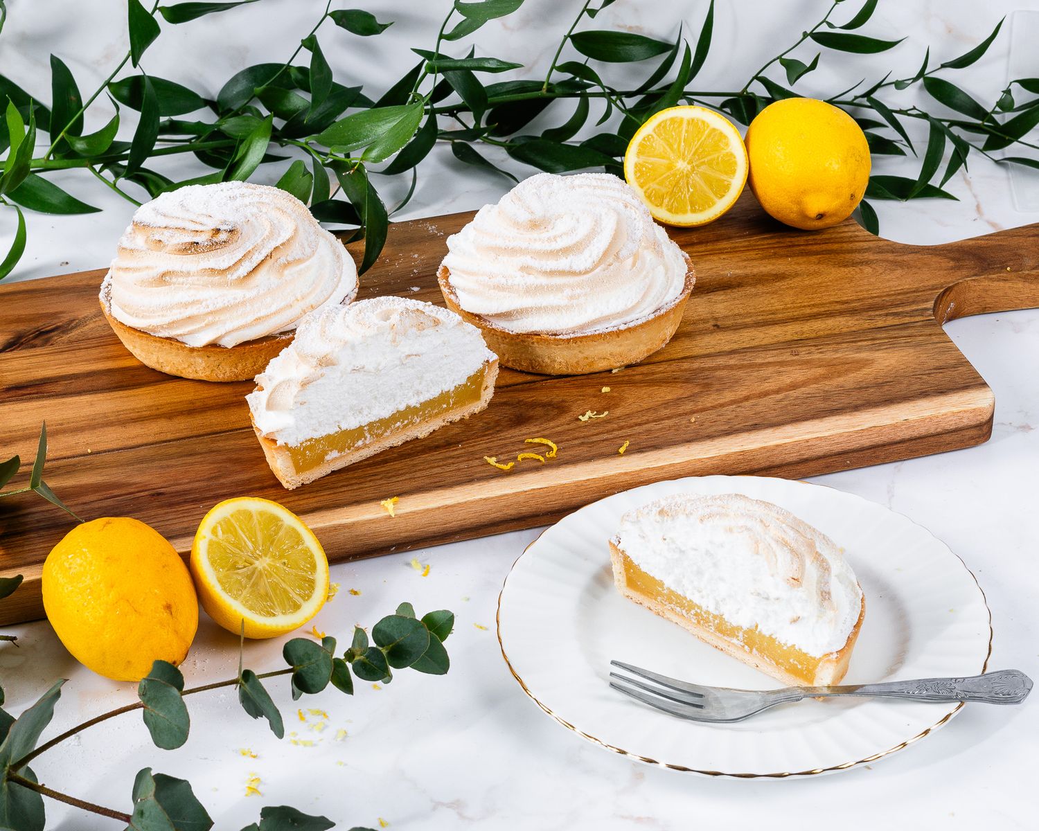 Three lemon meringue tarts on a wooden board with a sliced tart on a plate beside lemons.