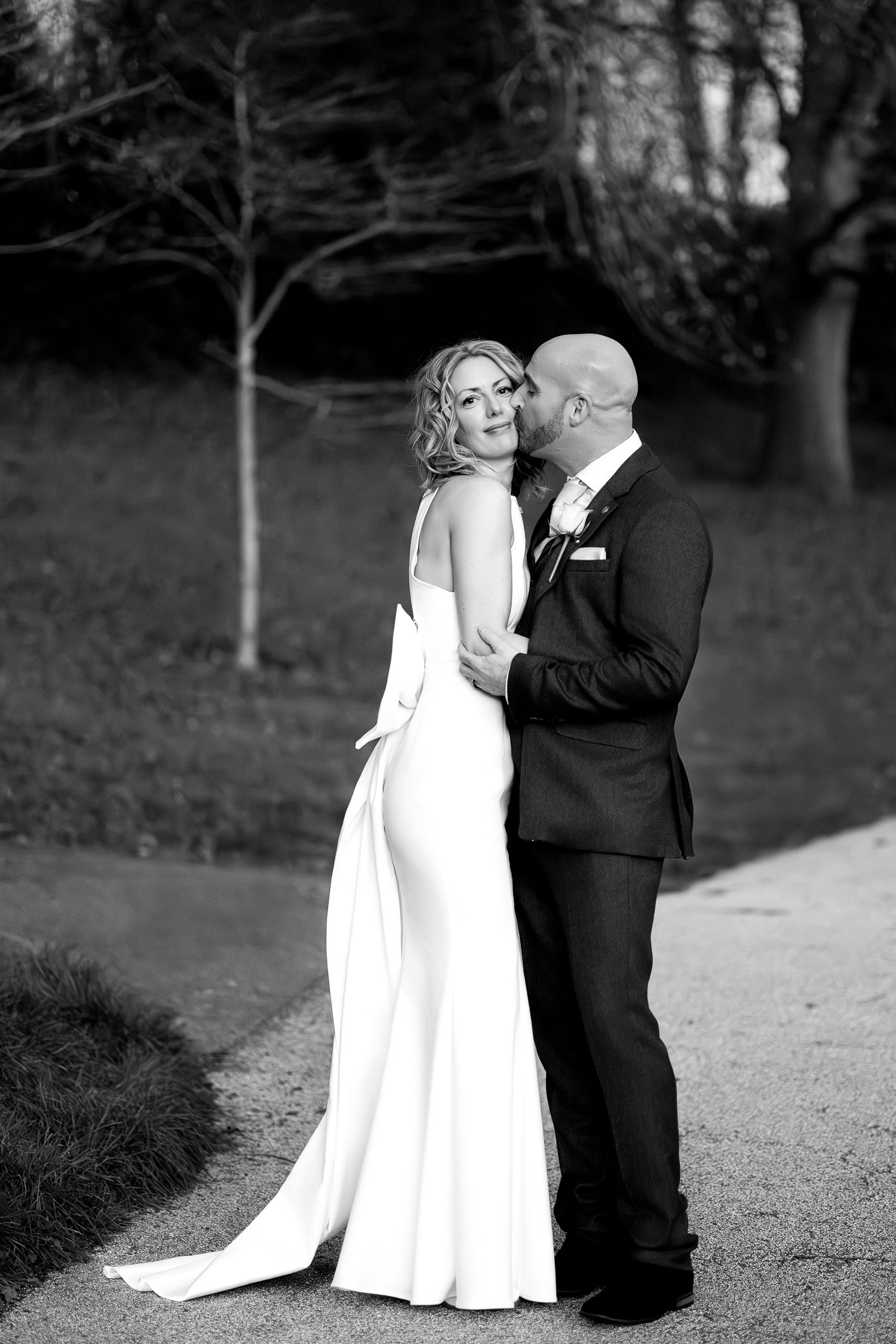 Bride and bald groom embrace on outdoor path in elegant black and white wedding portrait.