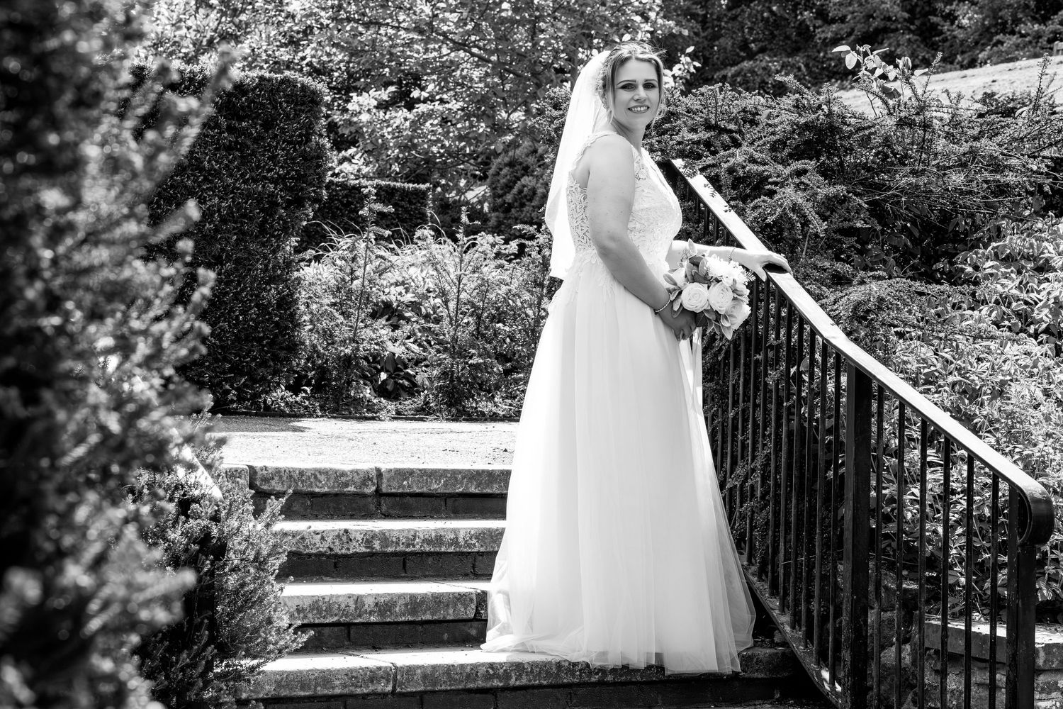 Bride in white gown and veil holding bouquet on stone steps in a lush garden, black and white wedding photo.