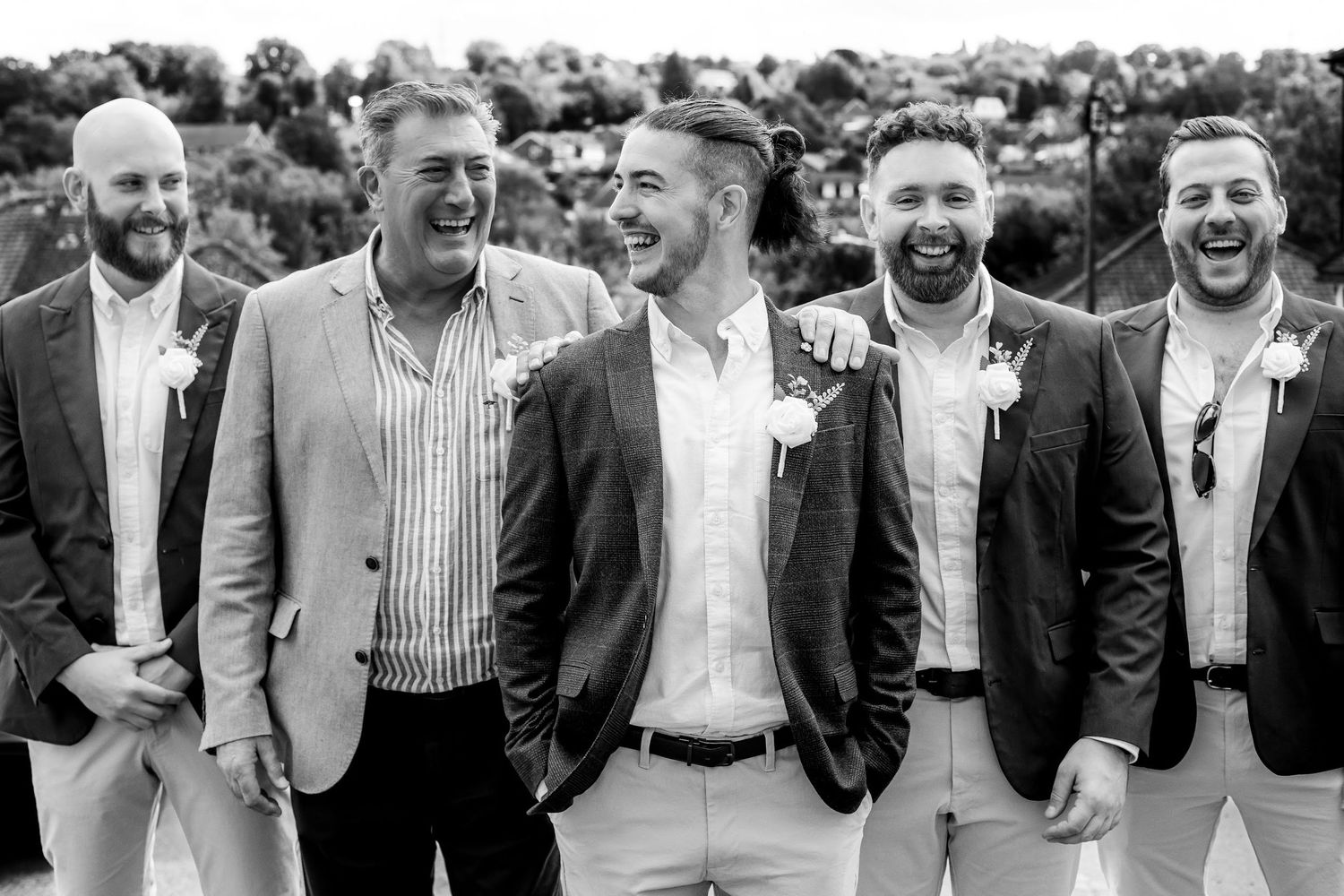 Black and white photo of five groomsmen laughing together outdoors at a wedding celebration.