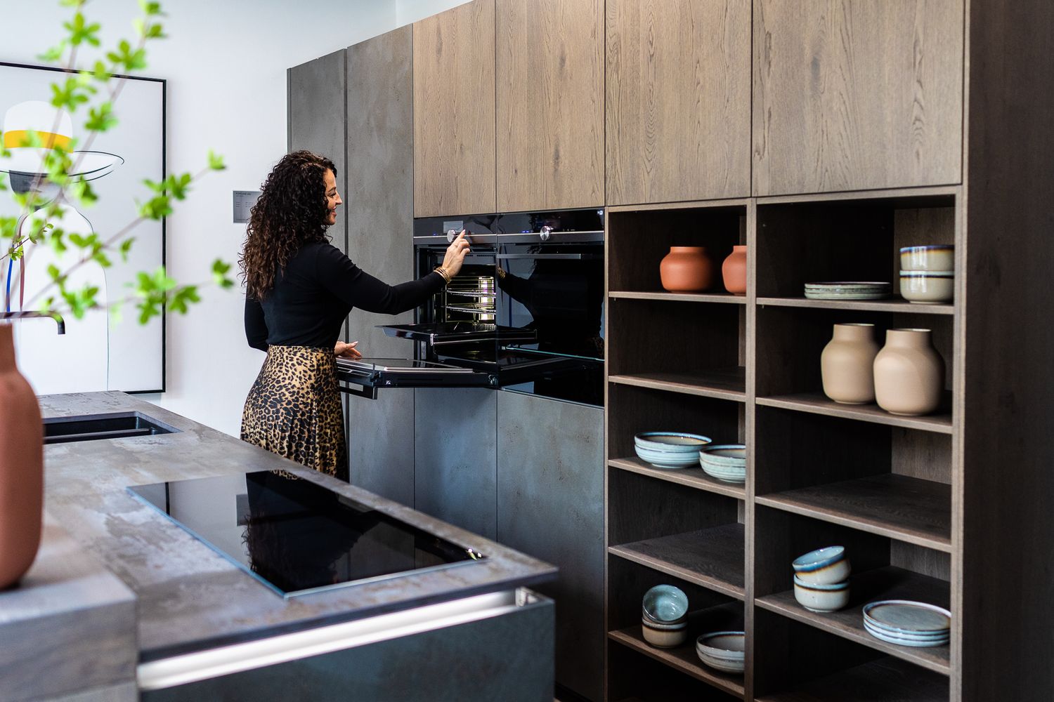 Woman using built-in oven in a modern Nolte kitchen with wood cabinets and open ceramic display shelving.