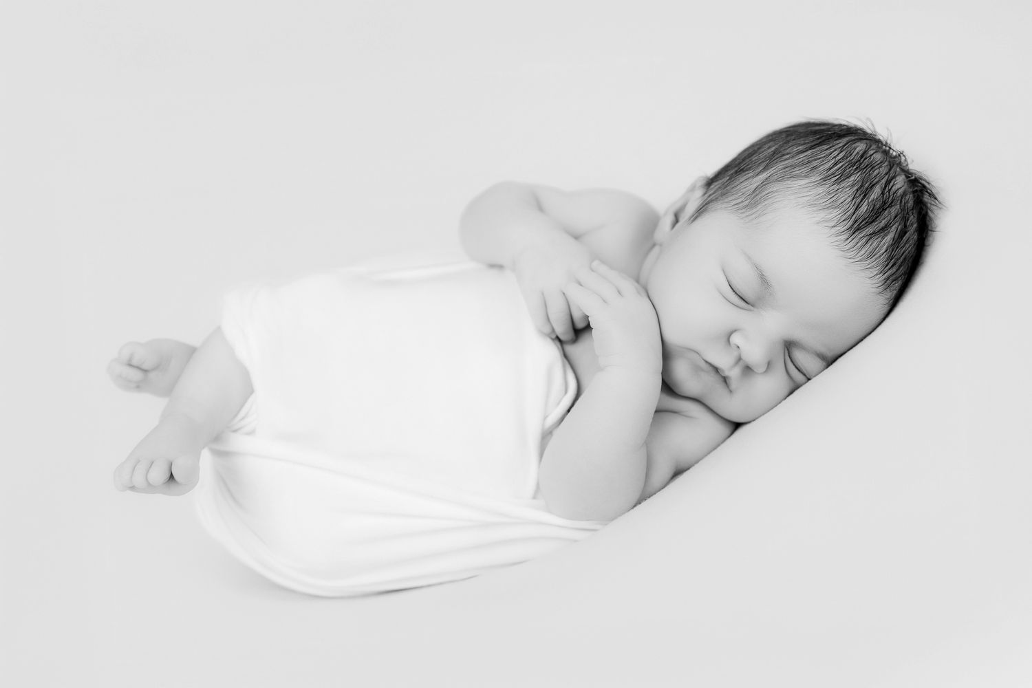 Sleeping newborn baby swaddled in white wrap, curled up peacefully, black and white photo.