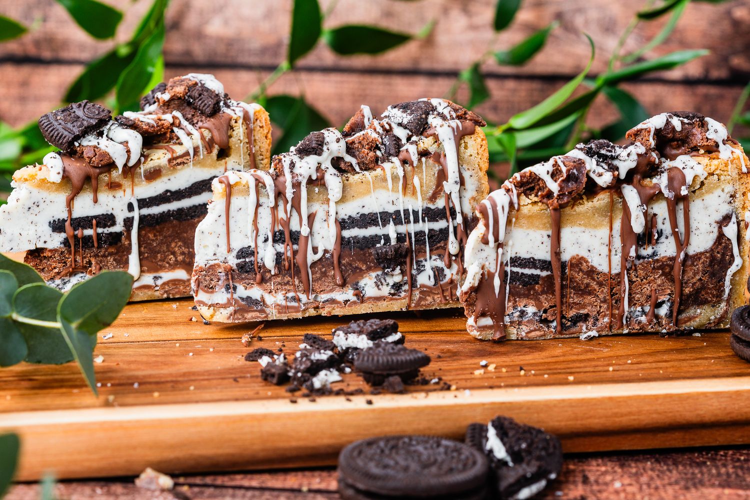 Oreo ice cream cake slices with chocolate drizzle on a wooden board with Oreo cookies.