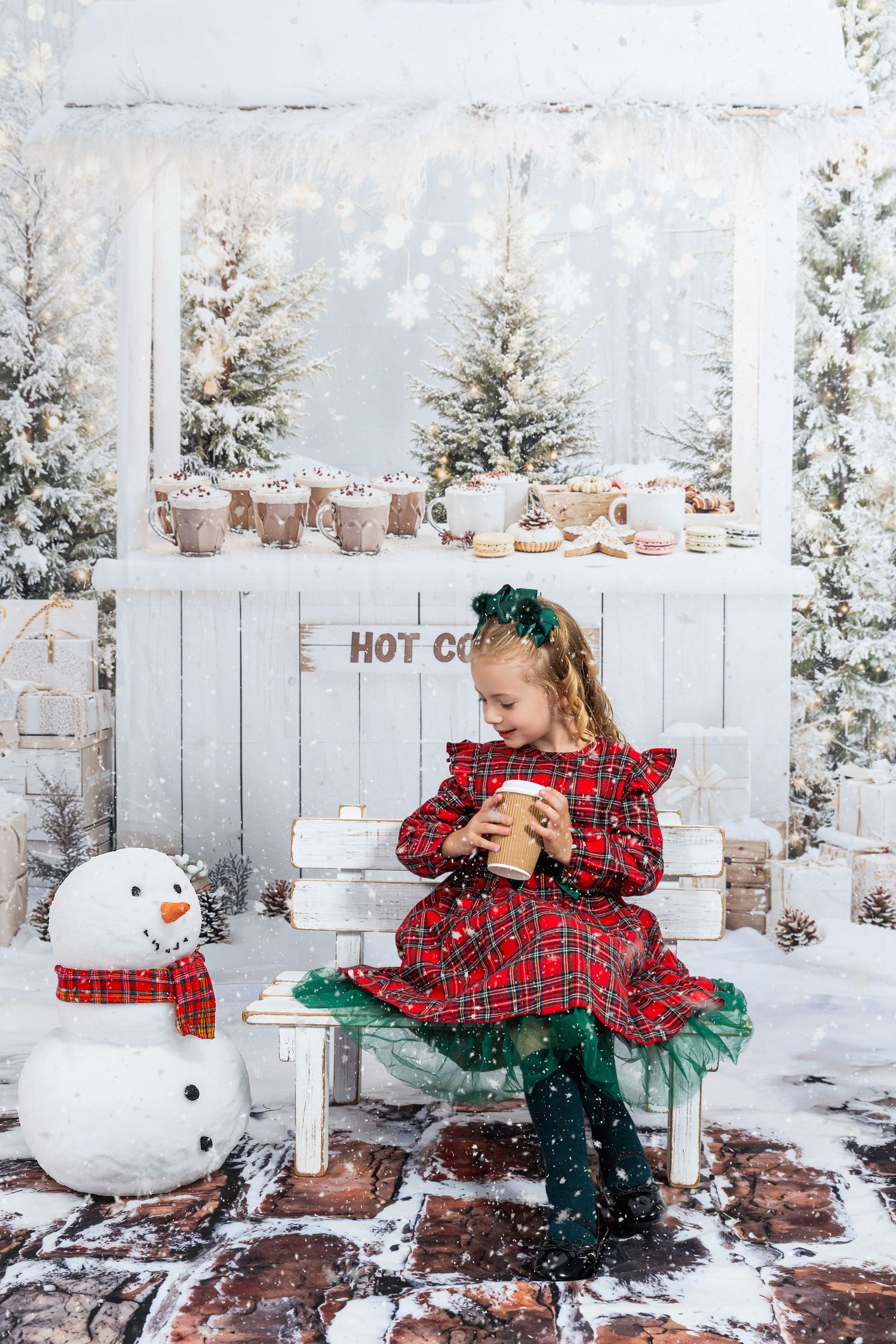 Young girl in red plaid dress holds hot cocoa cup beside snowman in festive Christmas winter wonderland photo setting.