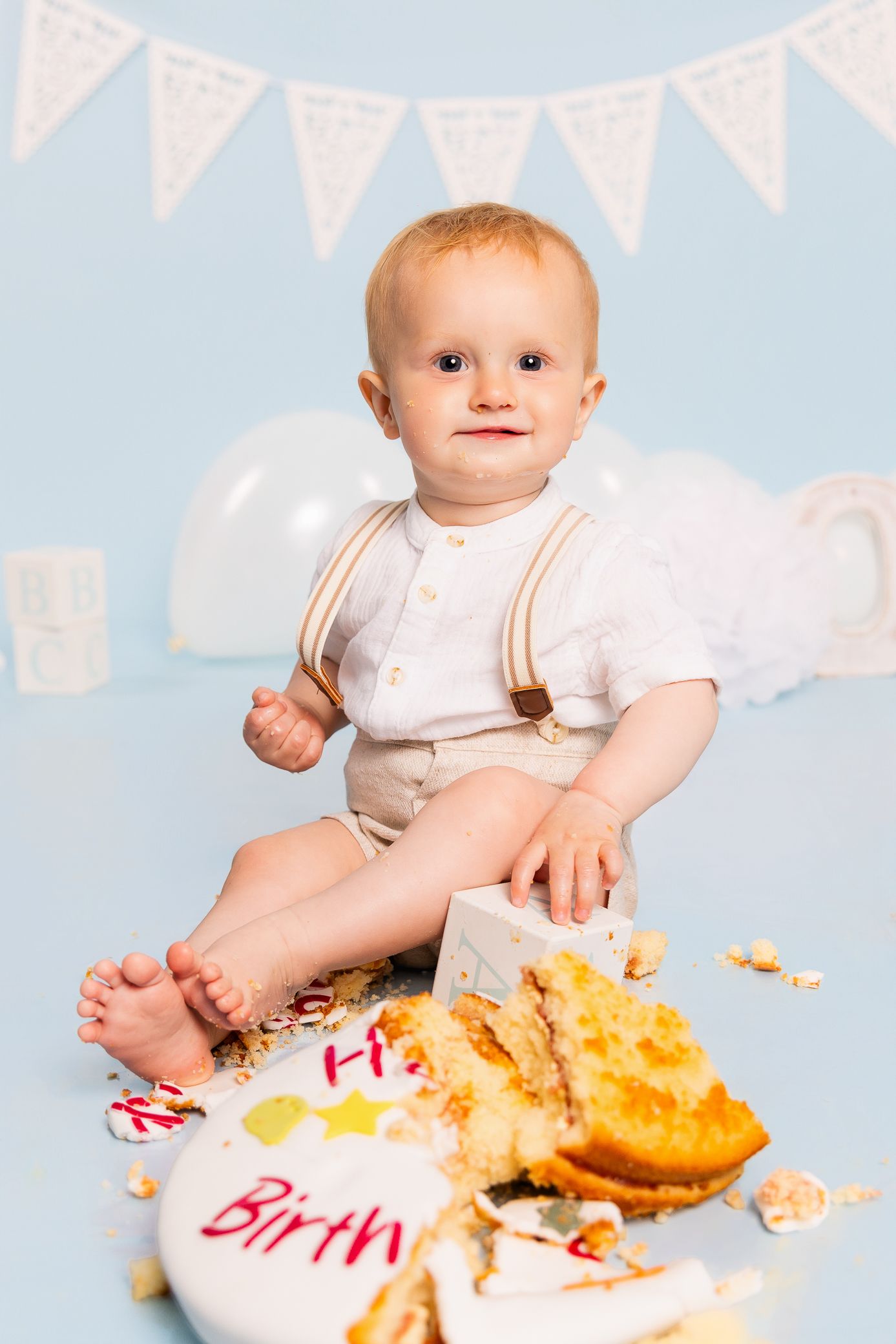 Adorable baby boy celebrating first birthday smashing cake, wearing suspenders, sitting on blue background with balloons.