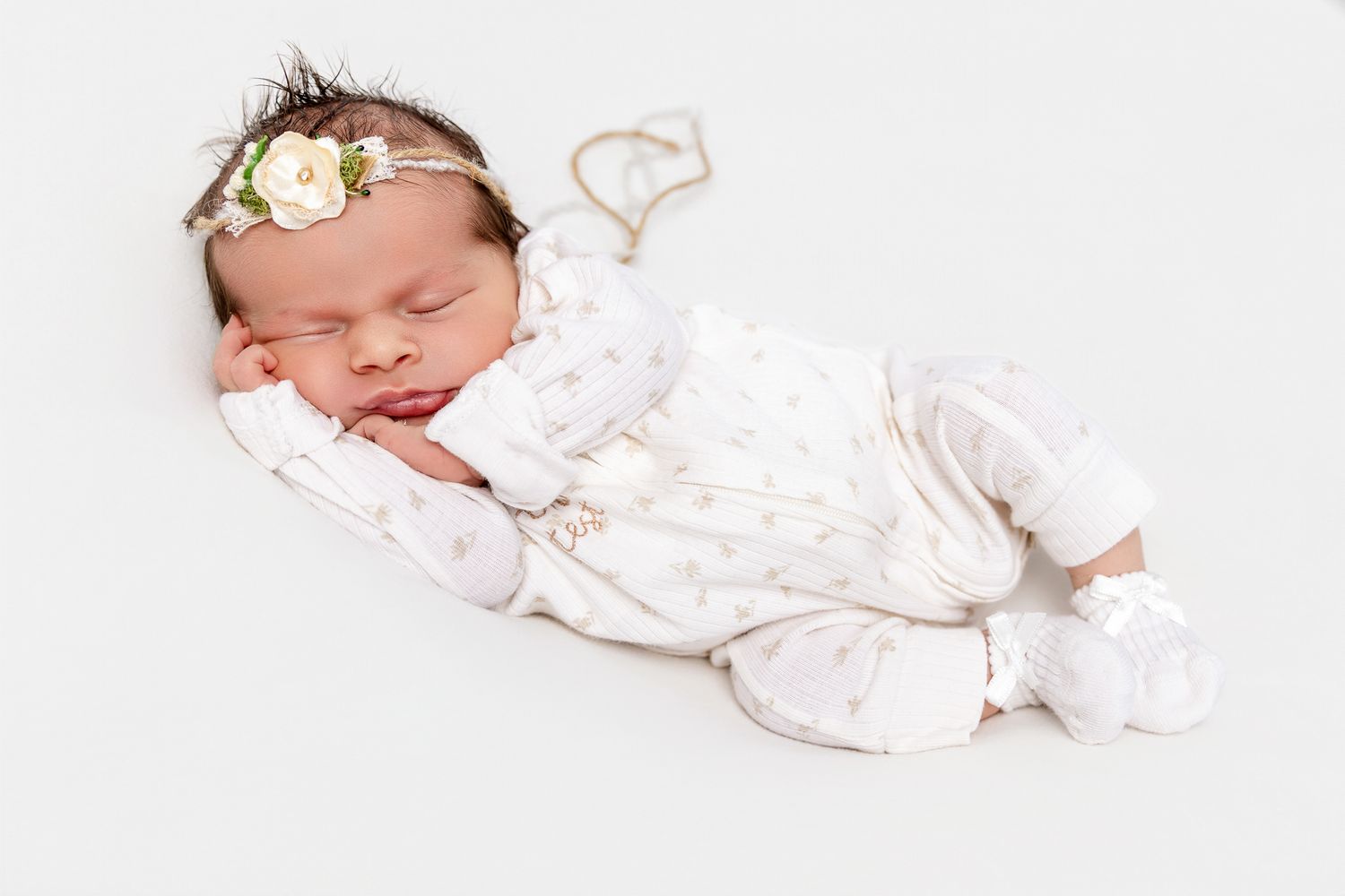 Sleeping newborn girl in white outfit with floral headband, lying on white background.