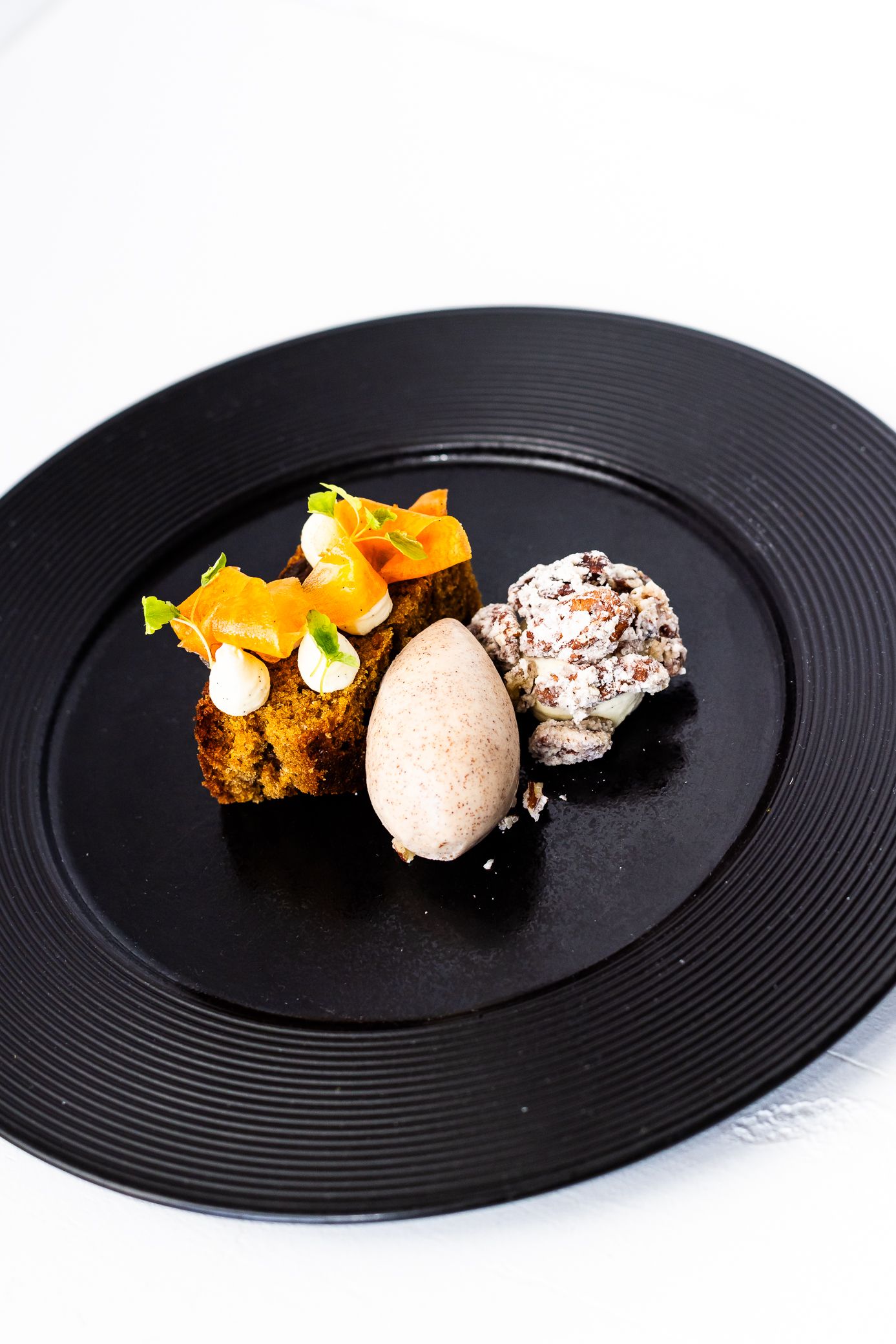 Fine dining dessert with chocolate ice cream, orange cake, and cream on black plate.
