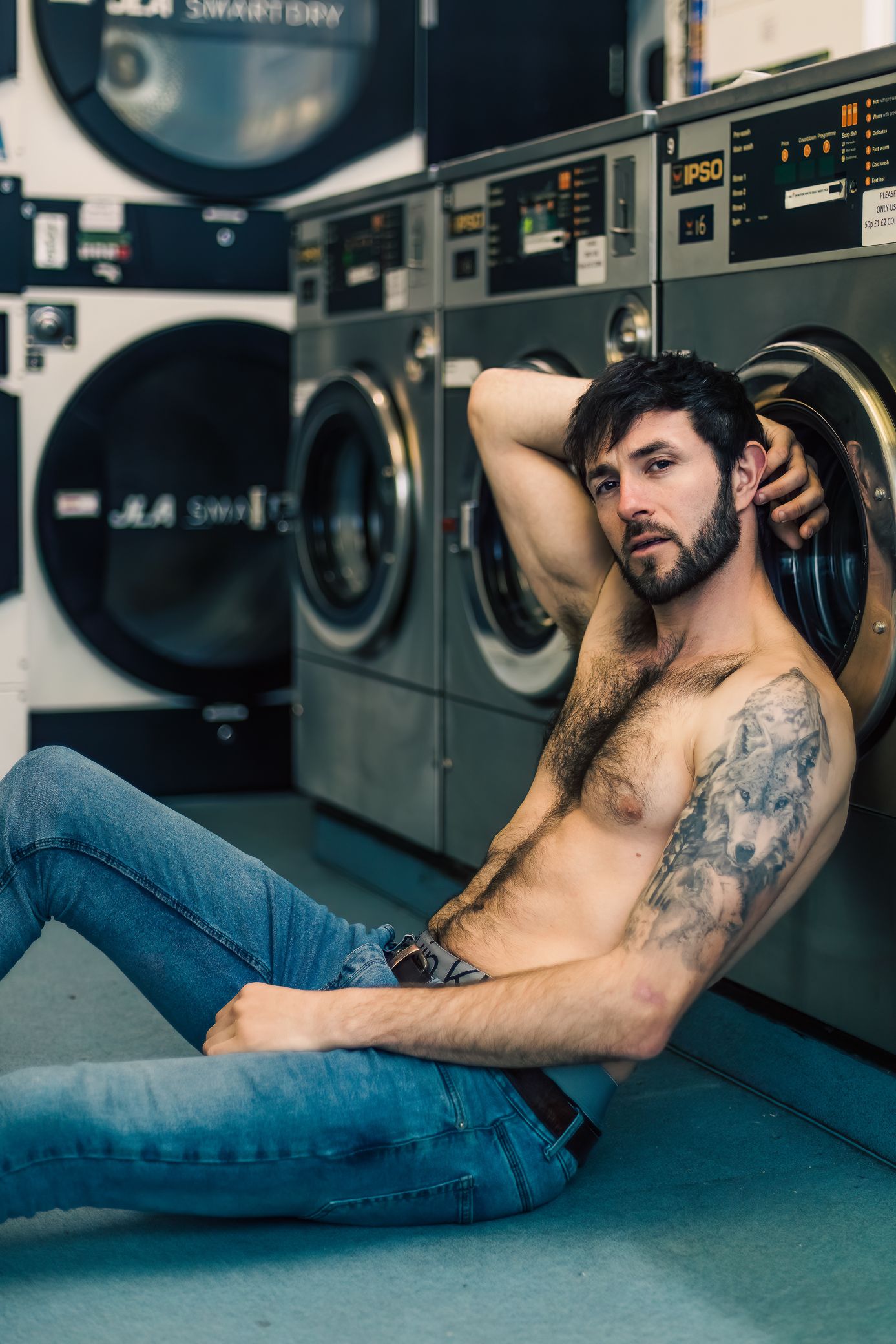 A tattooed shirtless man in jeans relaxes on the floor of a laundromat beside washing machines.