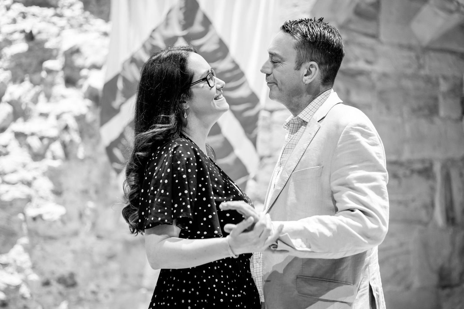 A couple shares a romantic moment, smiling at each other in a black and white outdoor photo.